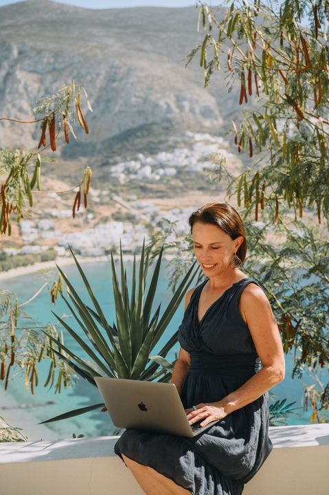 Woman sitting with laptop, smiling, overlooking a coastal village and turquoise water.