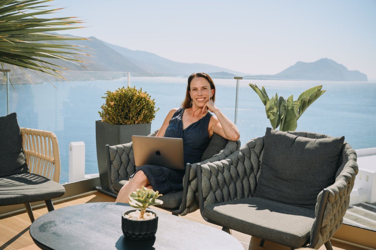 Woman working on a laptop on a balcony overlooking the sea, smiling.