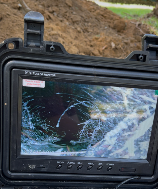 A handheld monitor displaying a live sewer camera feed, showing roots intruding into an underground pipe.