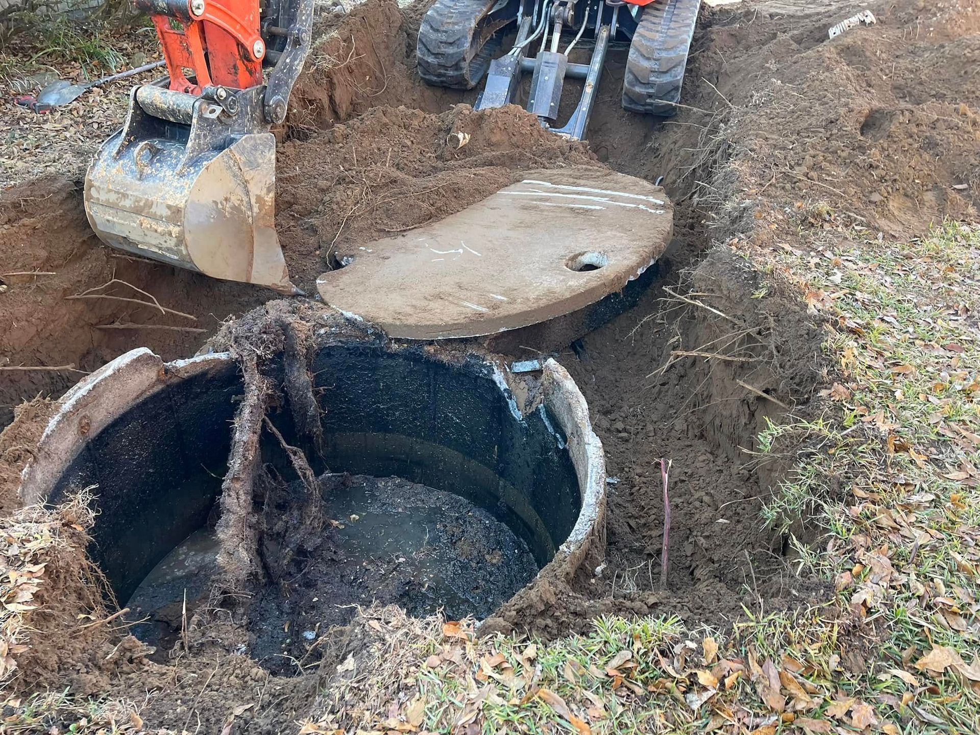 An orange excavator digs around an exposed septic tank and circular cover in a grass-covered yard.