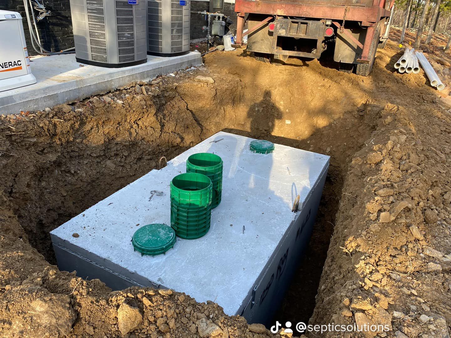 A large concrete septic tank sits in an excavated dirt pit, with several green risers and lids visible on top.