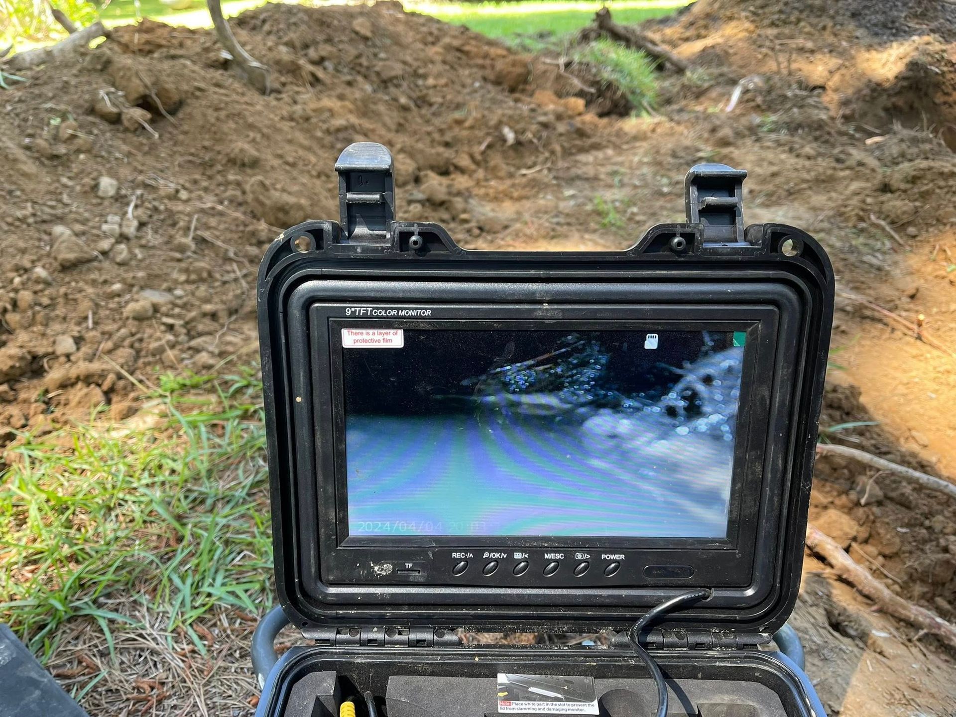 A monitor in a protective case displays a live feed of a dark pipe interior, positioned in an outdoor trench with dirt.