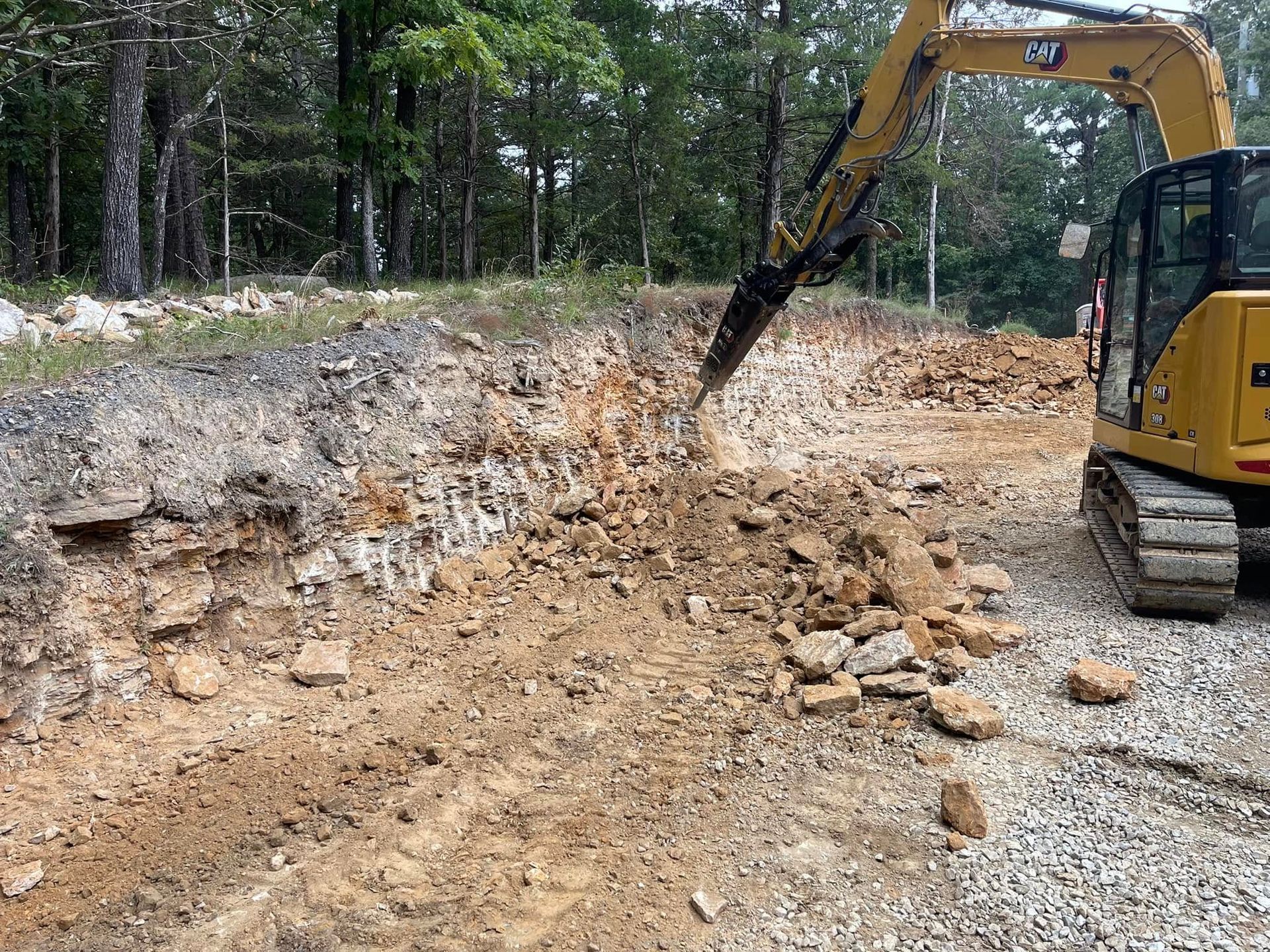 A yellow CAT excavator uses a hydraulic hammer attachment to break up rocky soil on a wooded construction site.