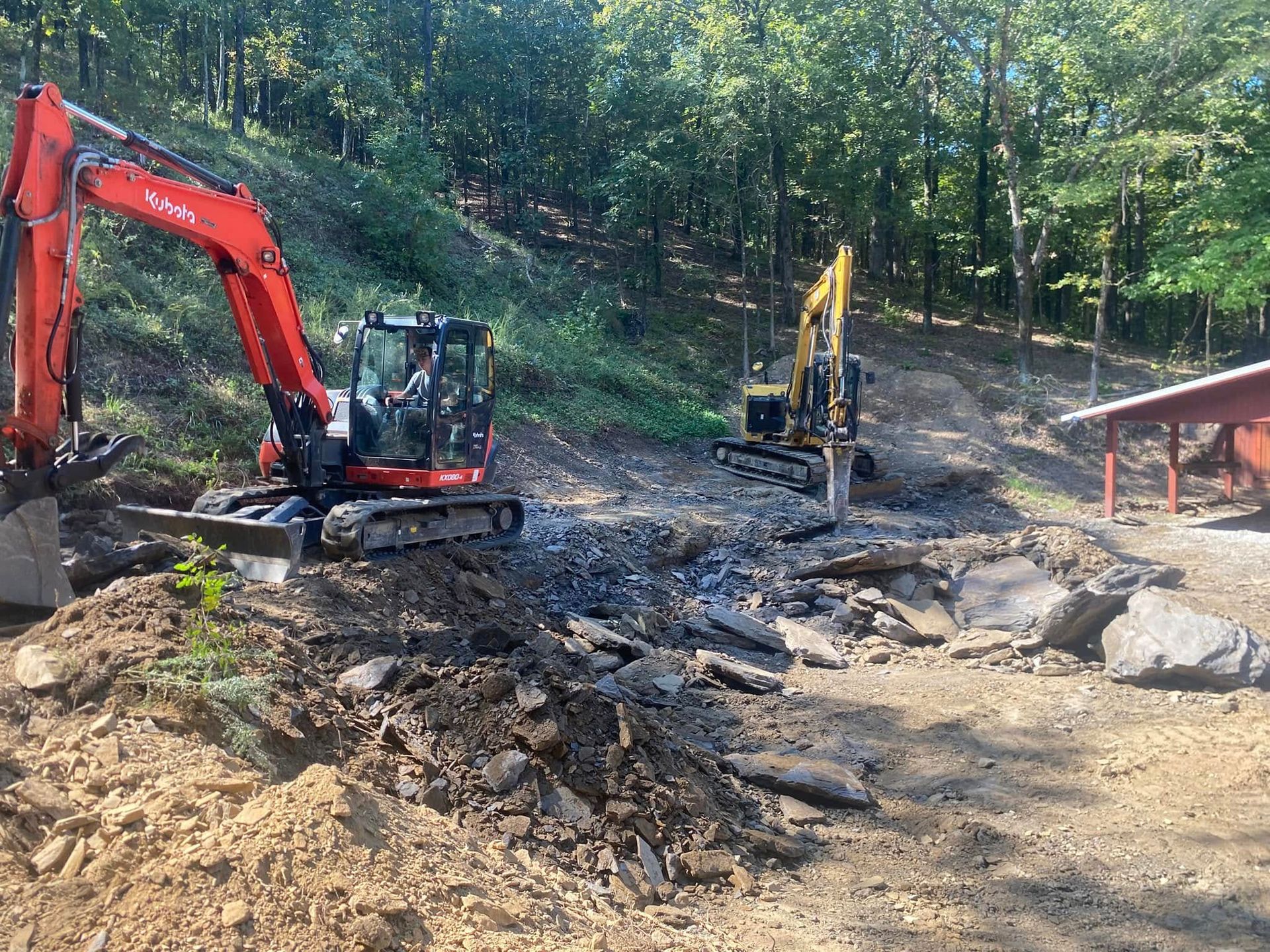 Two excavators working on a rocky, dirt-filled clearing near a forest and a small wooden structure.