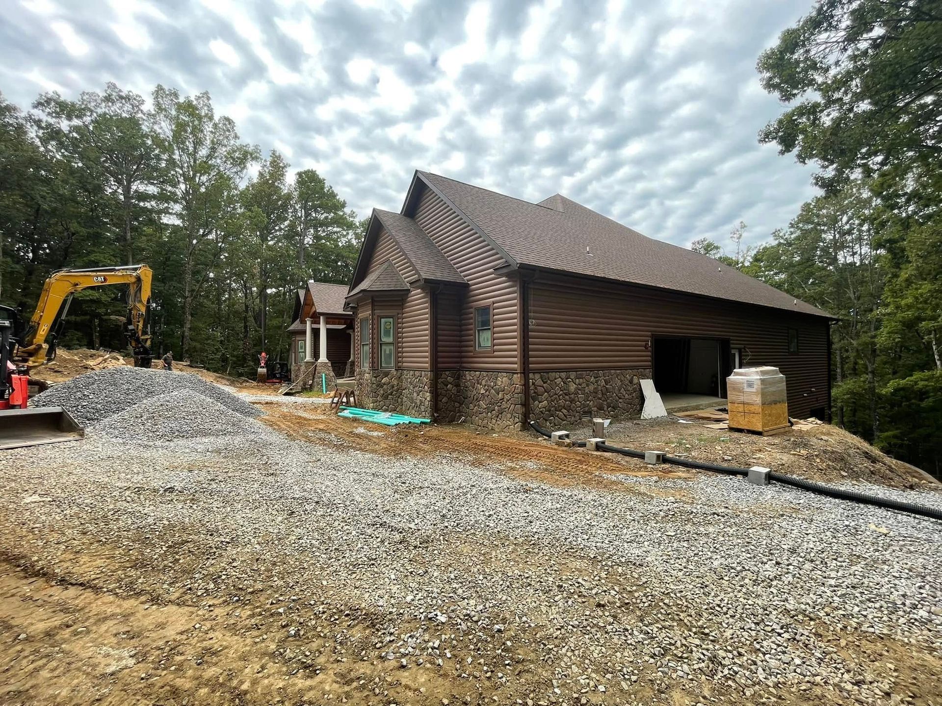A house under construction with brown siding and stone base, next to a gravel driveway and an excavator in a wooded area.