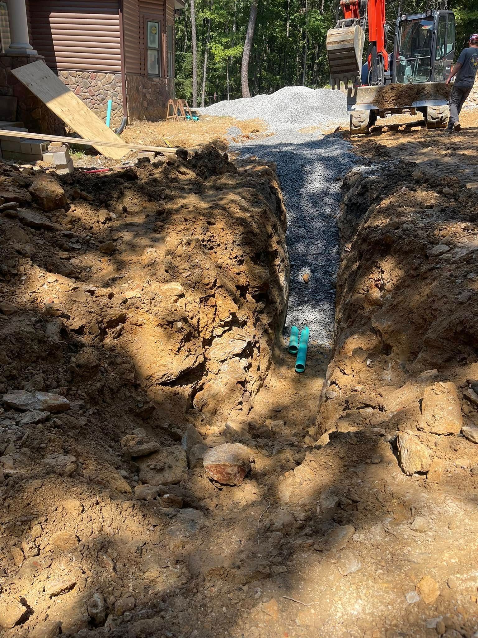 A deep dirt trench leads toward an orange excavator parked near a house under construction with stone-covered drainage.