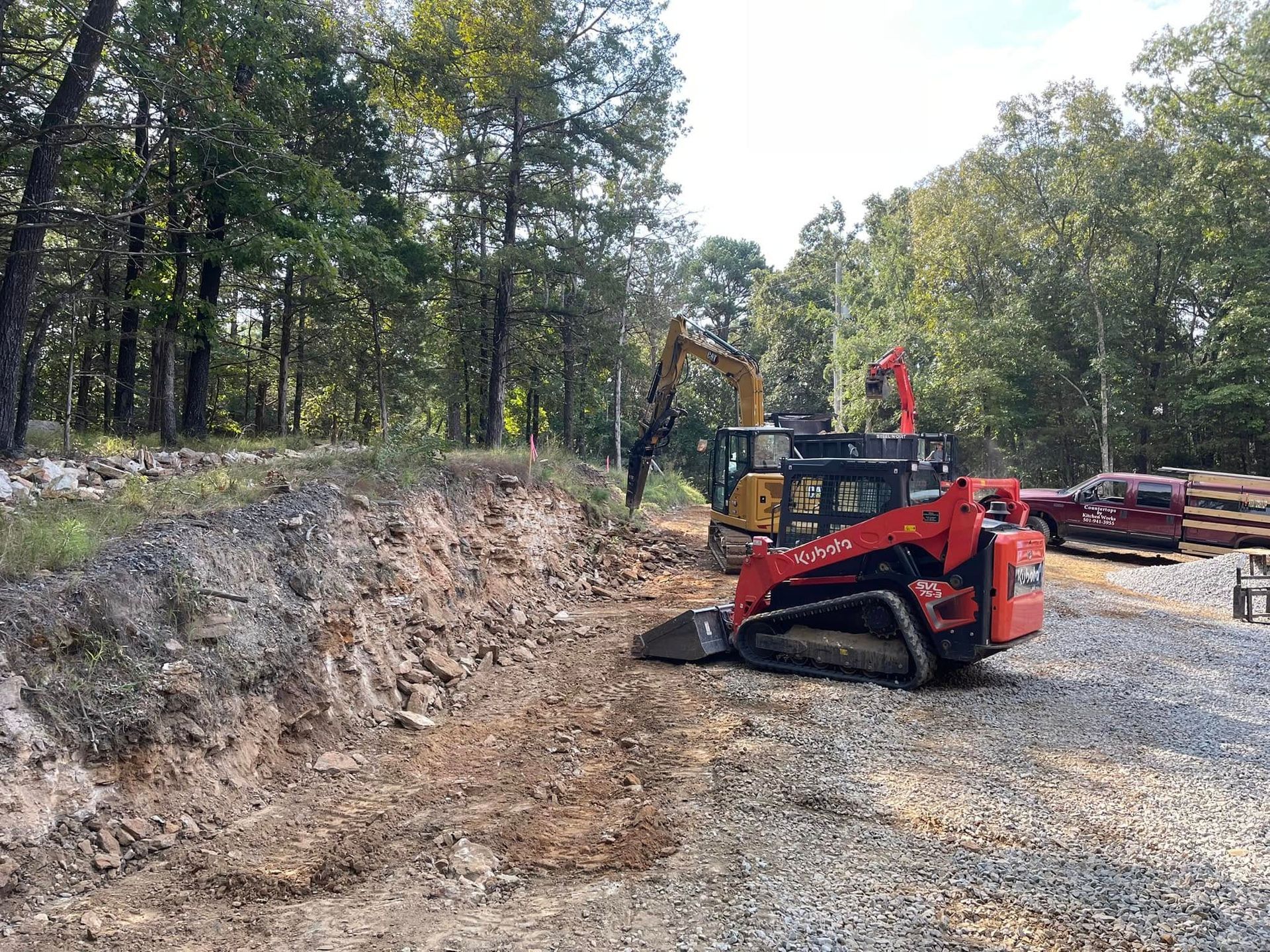 A red skid steer loader and a yellow excavator work on a rocky, cleared site near a forest and a parked pickup truck.