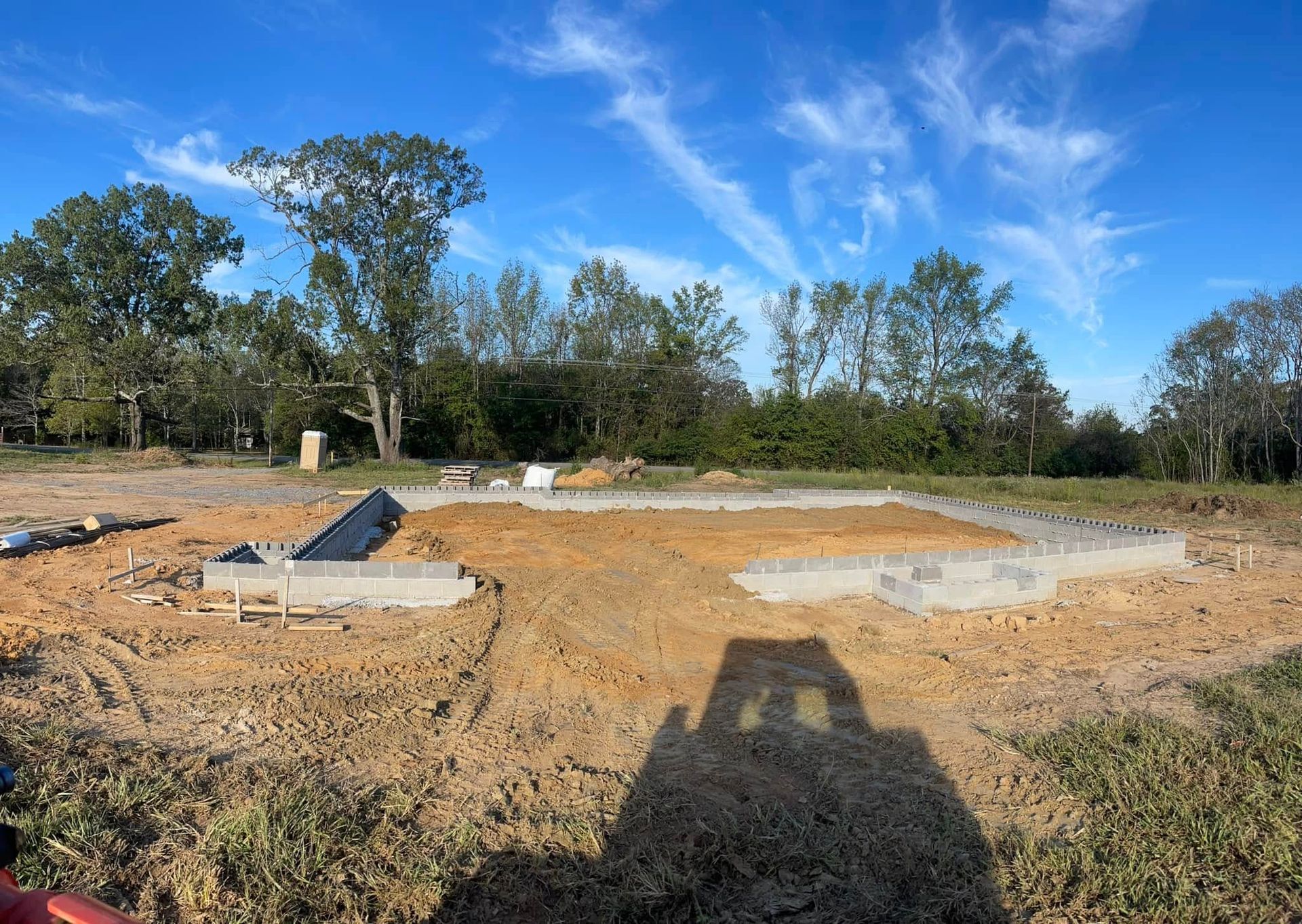 A construction site shows a partially built concrete foundation surrounded by dirt, with a treeline under a blue sky.