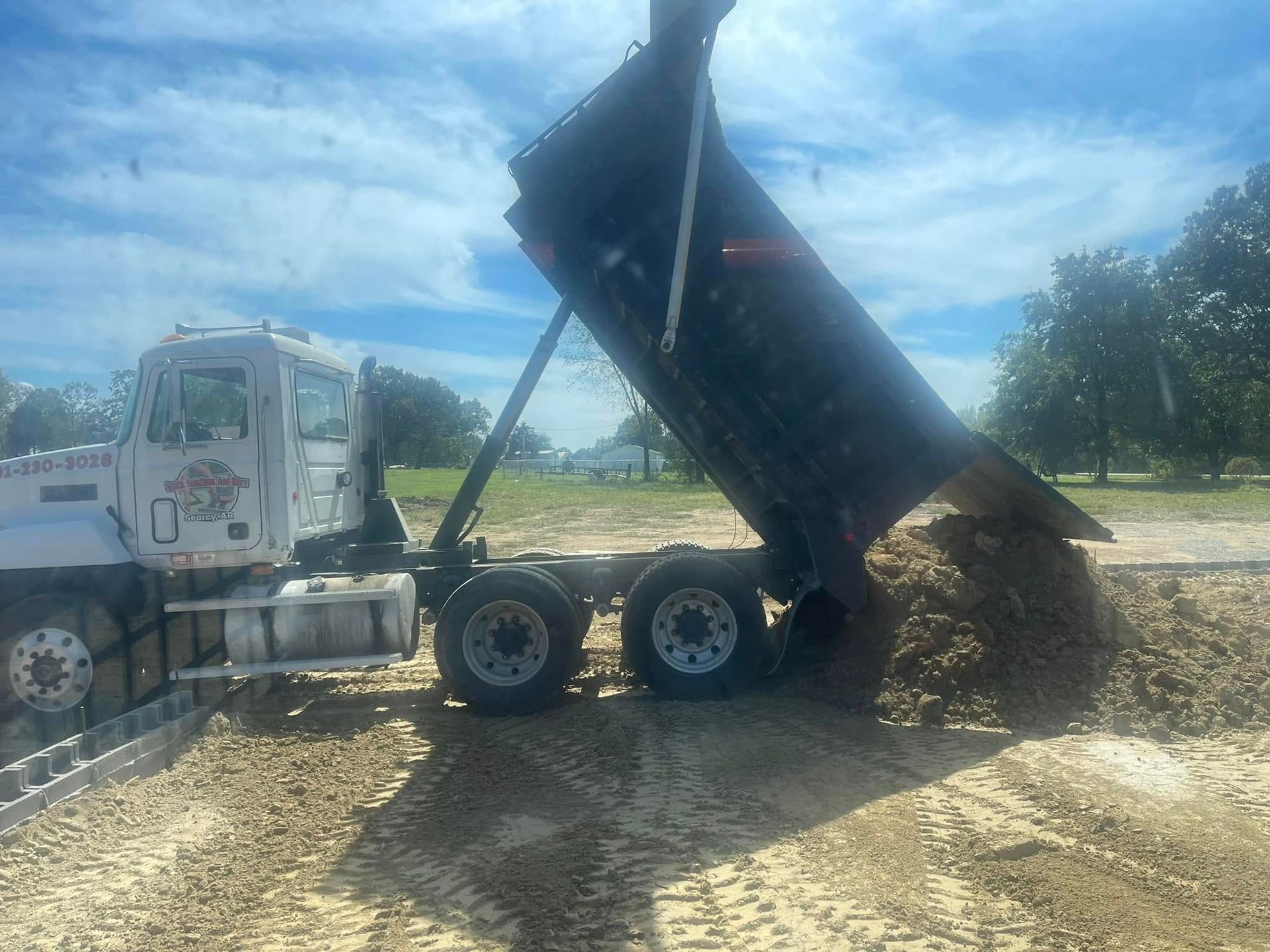 A white dump truck on a dirt lot with its bed raised, emptying a pile of soil onto the ground under a sunny sky.