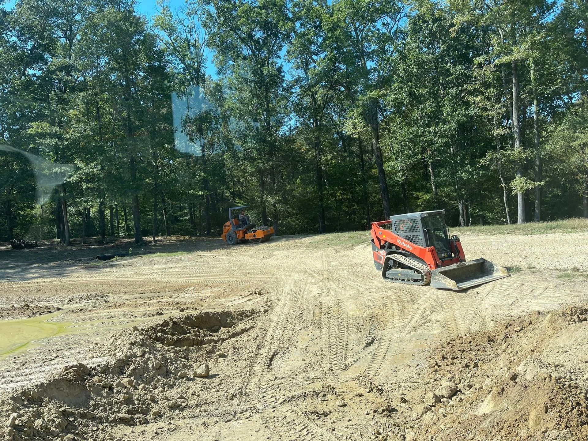 An orange skid-steer loader and a yellow roller on a dirt construction site in front of a forest.