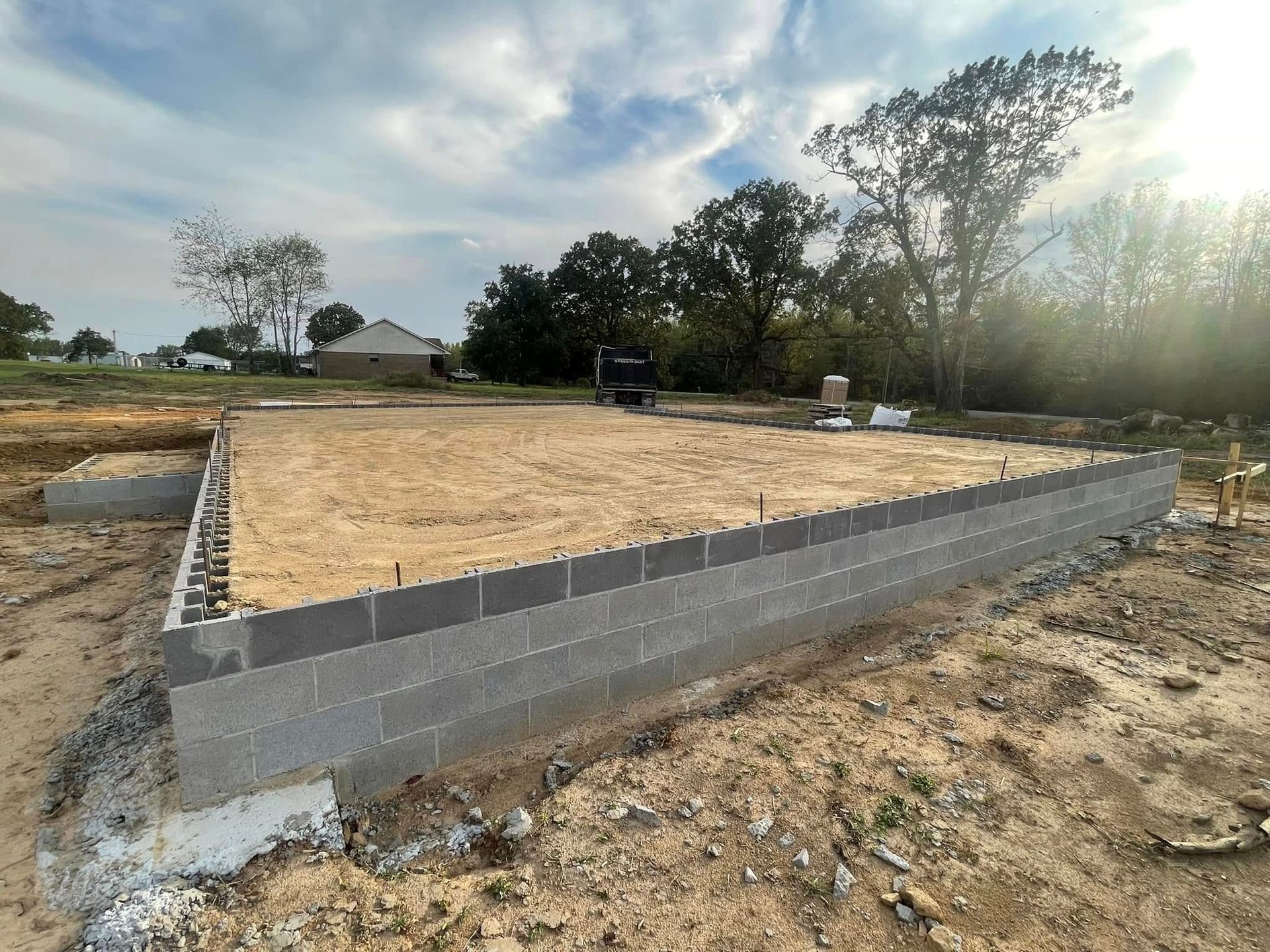 A building foundation made of grey concrete blocks surrounds a sand-filled plot on a construction site with trees.