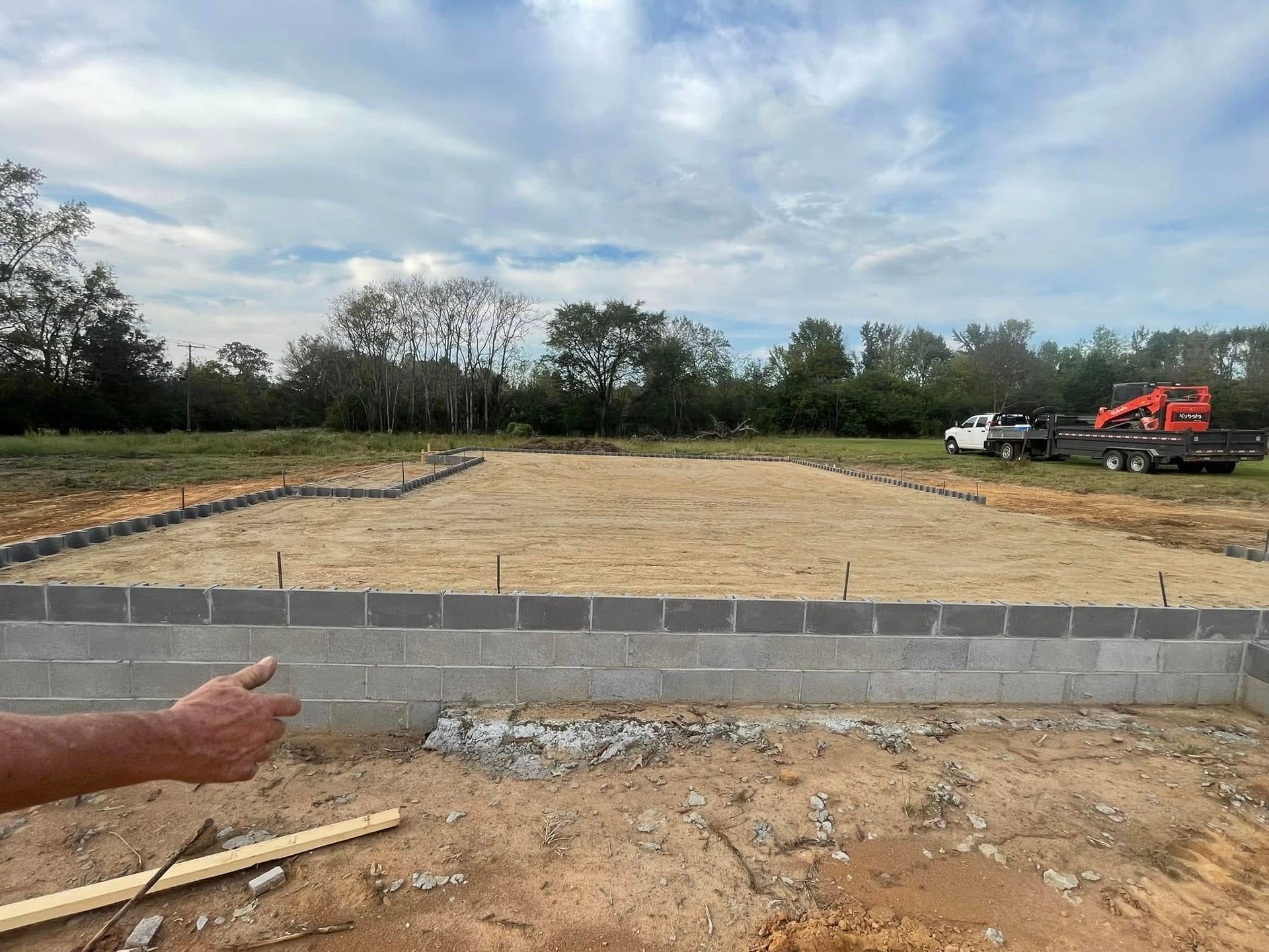 A construction site showing a concrete block foundation wall around a dirt pad under a partly cloudy sky.