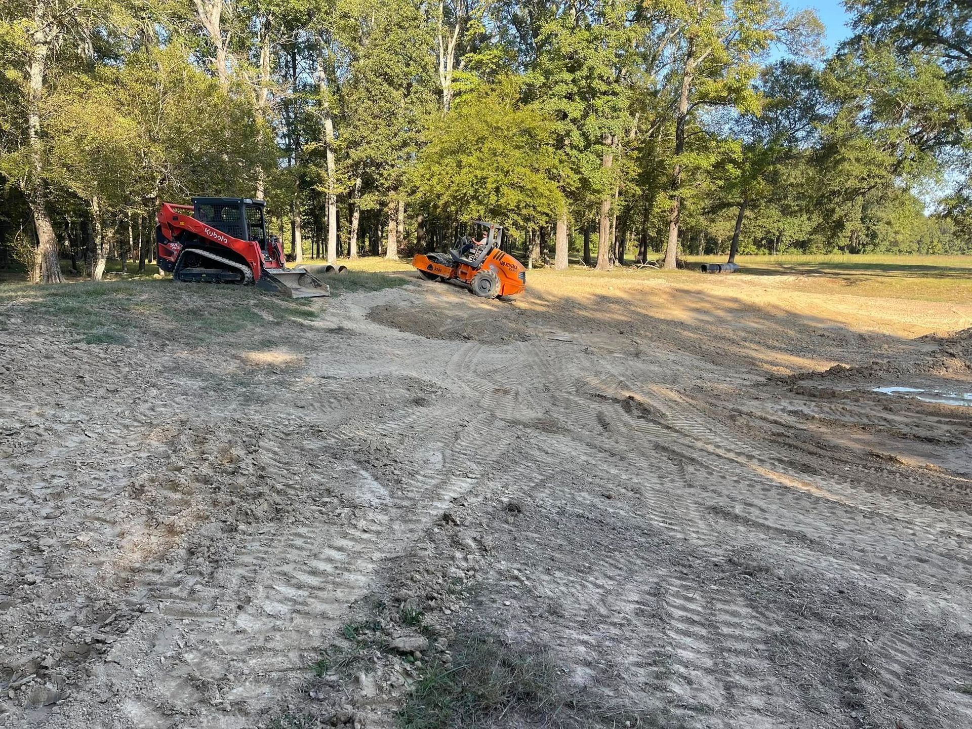 Two orange construction vehicles level dirt on a cleared lot with trees in the background under a blue sky.