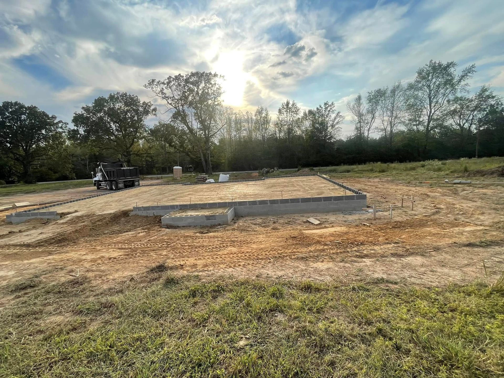 A concrete foundation for a new house under construction in a grassy field under a bright sunlit sky.