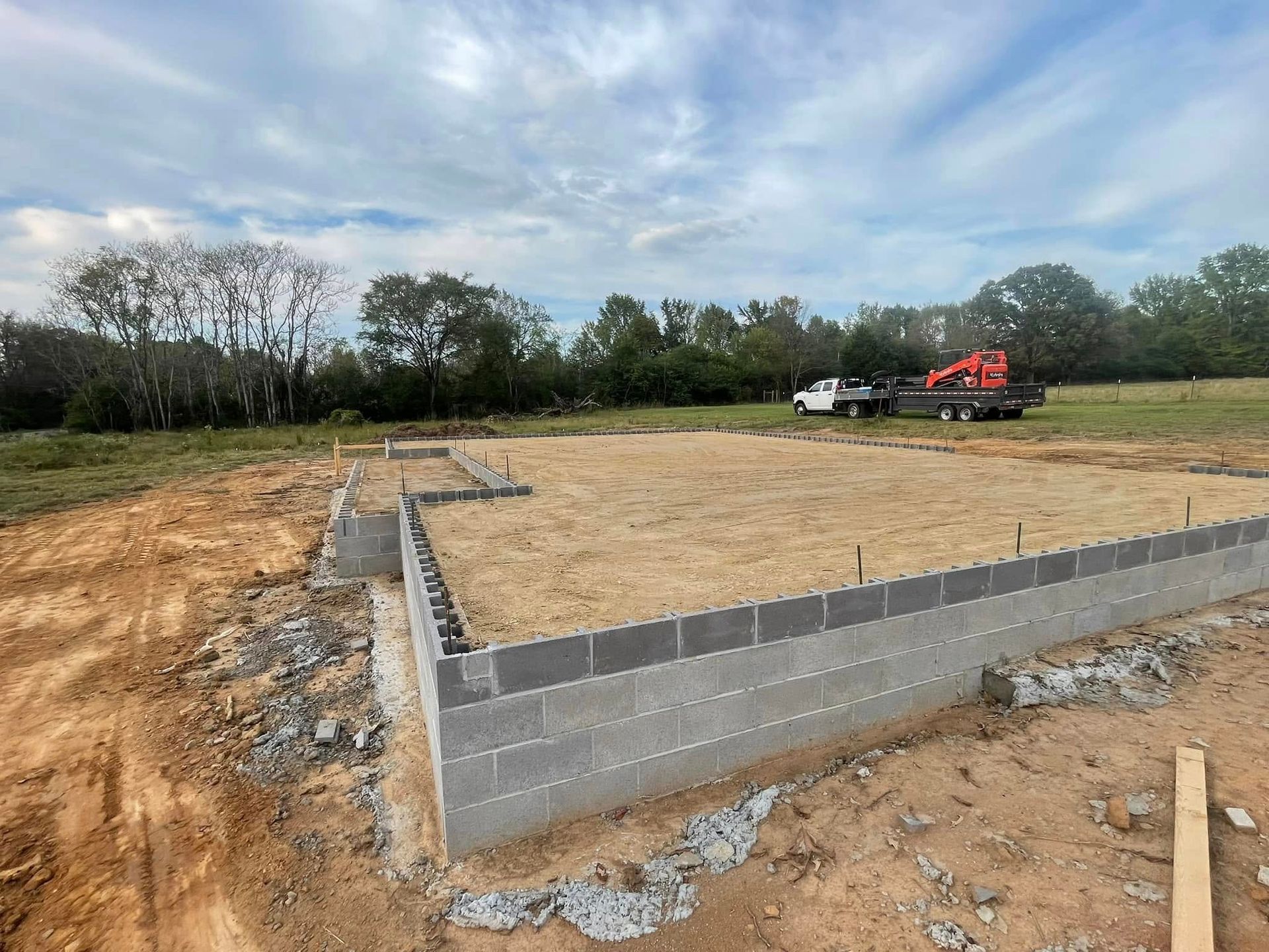 A cinder block foundation for a building under construction, set in a rural, grassy landscape with a truck in the distance.