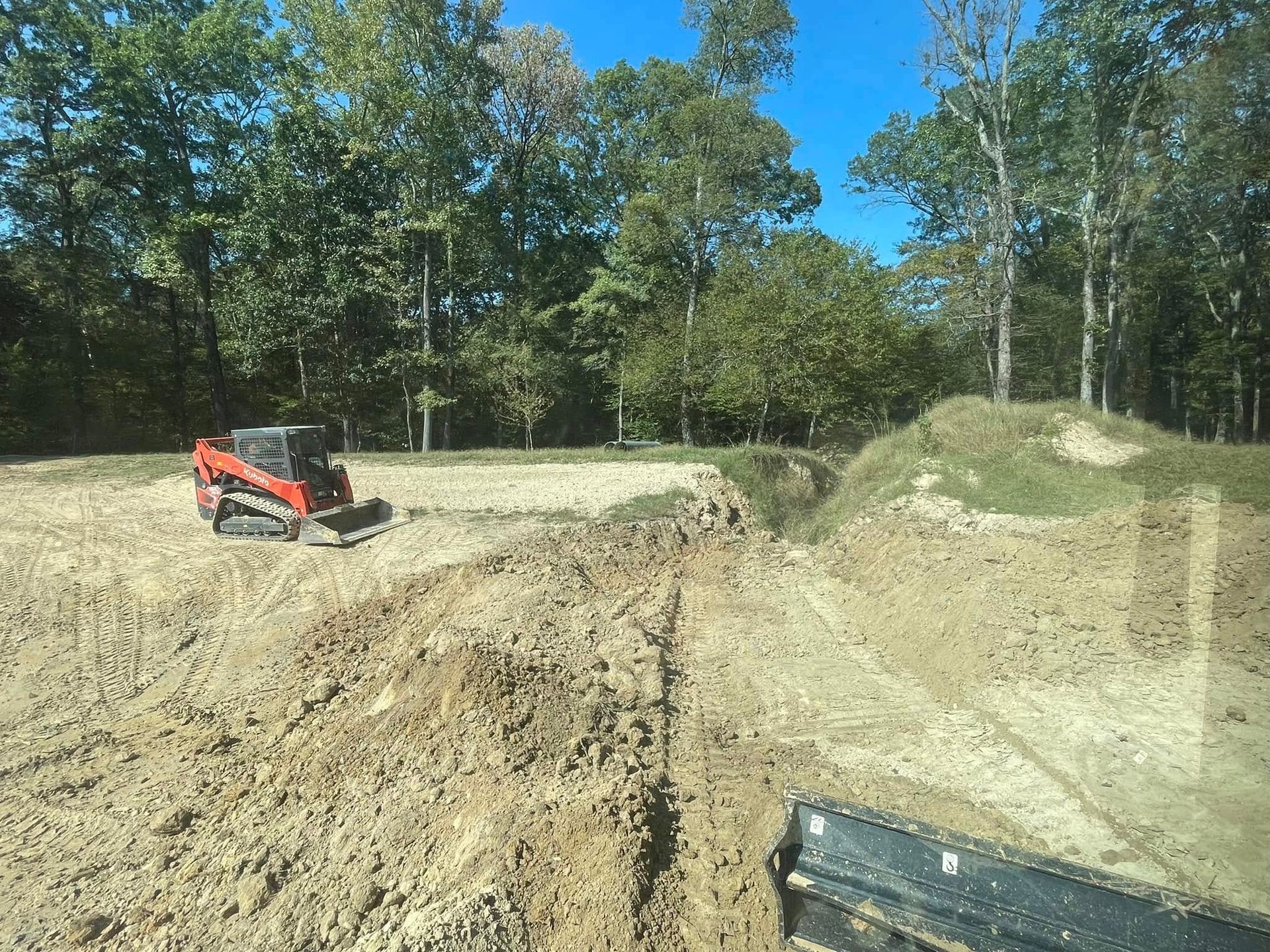 A red skid steer loader working on a cleared dirt construction site bordered by a line of trees under a clear blue sky.