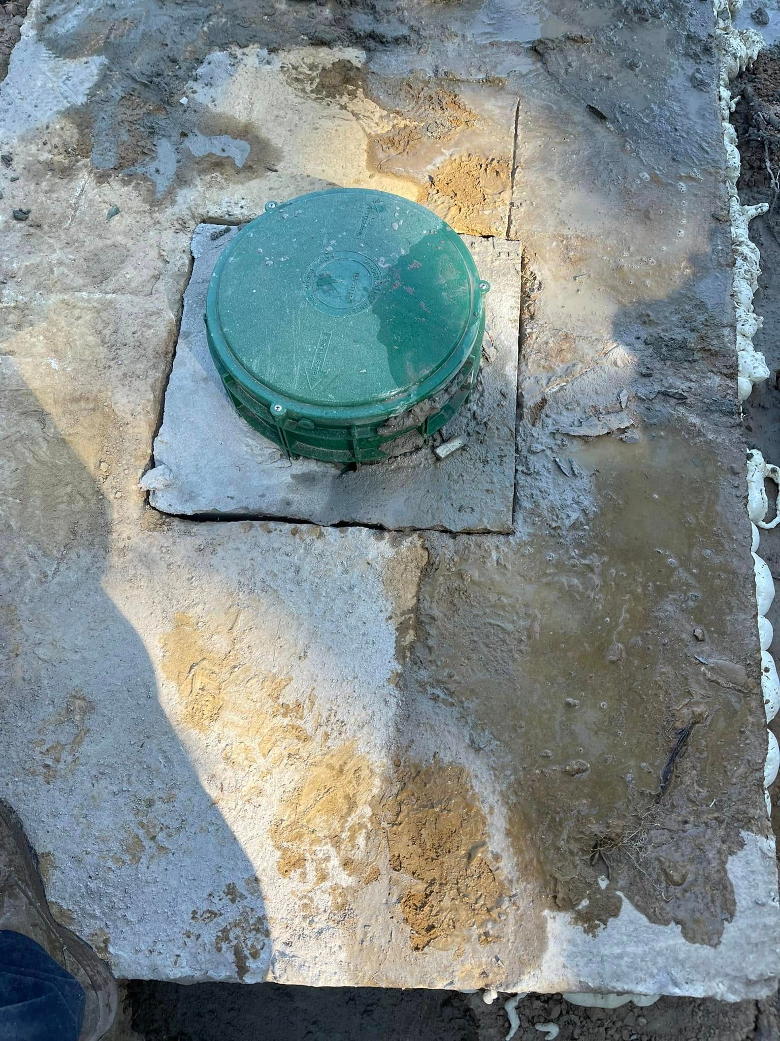 A circular green plastic access lid set into a weathered, light-colored concrete slab on the ground.
