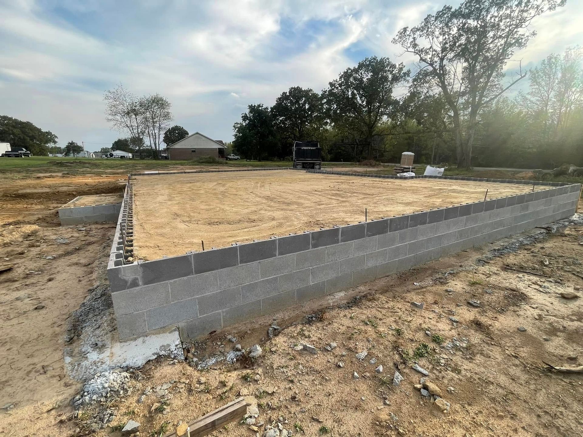 A rectangular cinder block foundation wall surrounds a plot filled with sand at a construction site under a partly cloudy sky.