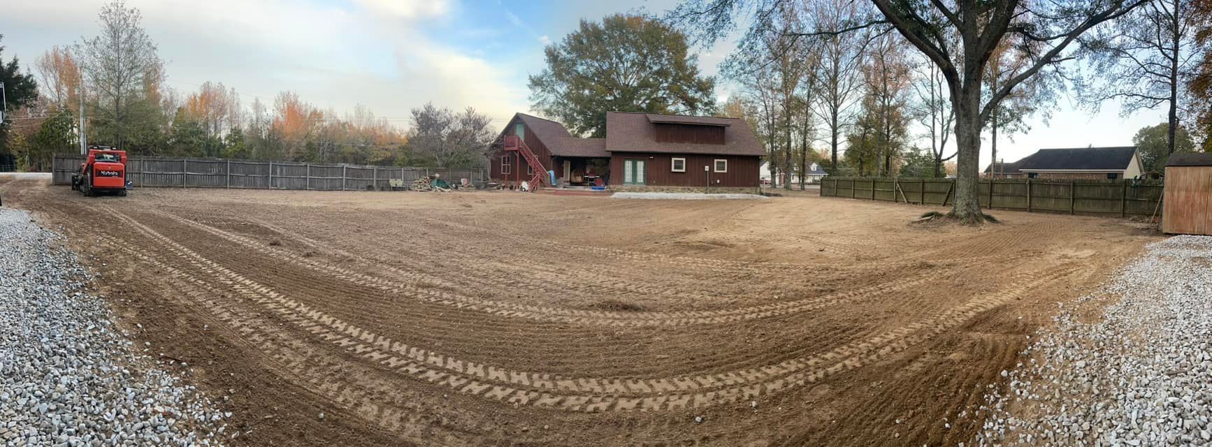 A wide-angle view of a large, bare dirt yard being leveled by a small red construction roller in front of a brown house.