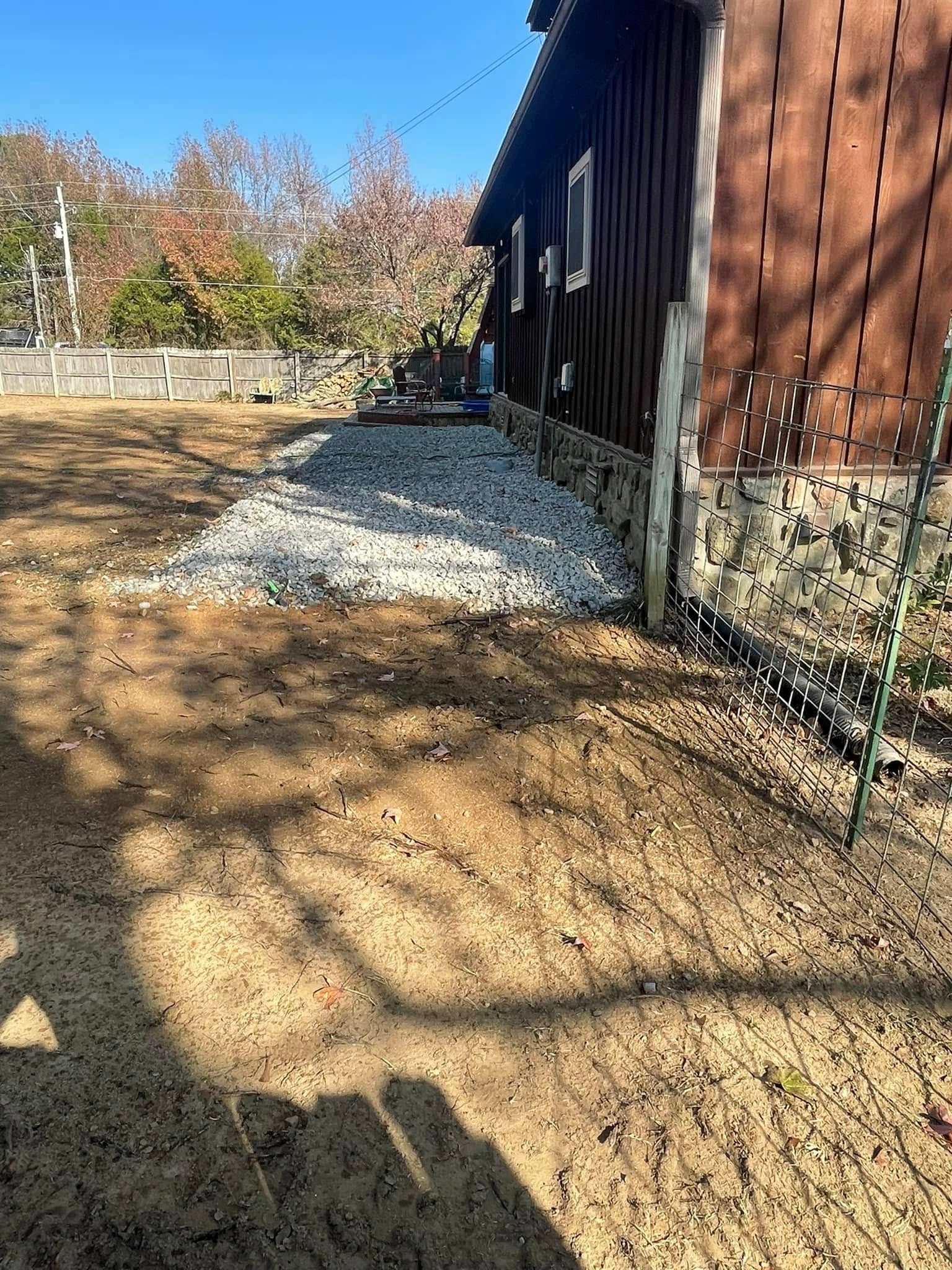 A gravel border runs along the stone foundation of a brown wooden building in a dry, sunny yard.