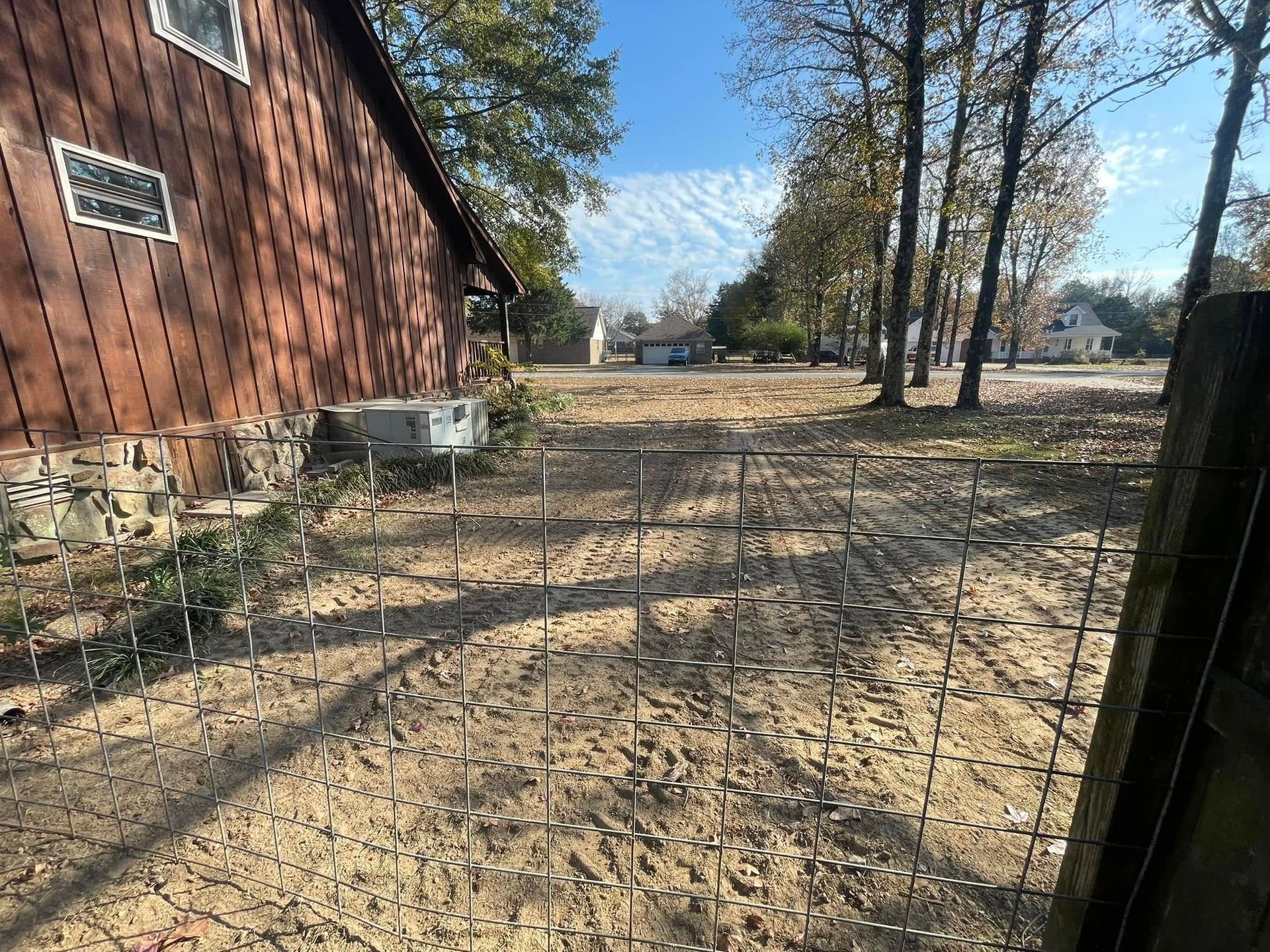 A red house exterior with a stone foundation beside a fenced-in, sandy lot with trees under a clear blue sky.