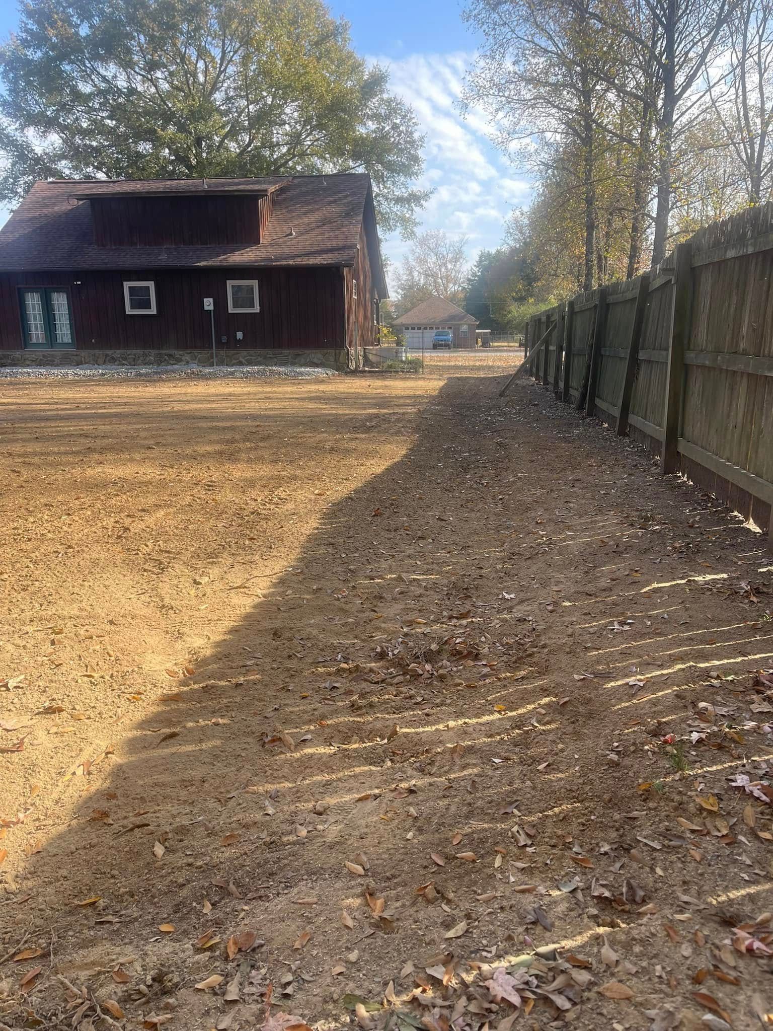 A red barn stands at the end of a leaf-covered yard beside a tall wooden fence under a sunny, blue sky.