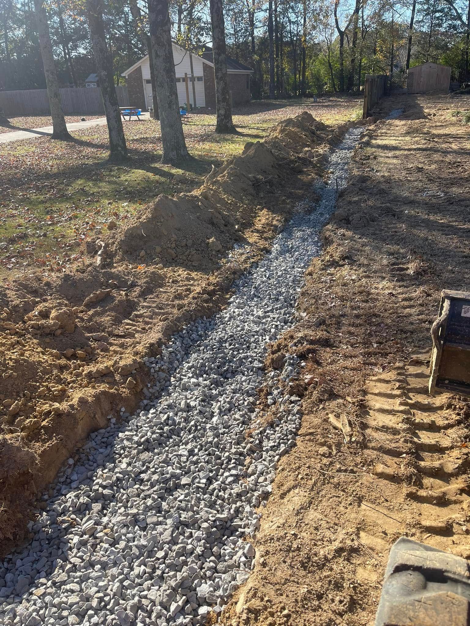 A gravel-filled drainage trench dug into a dirt yard, leading toward a house in the background.
