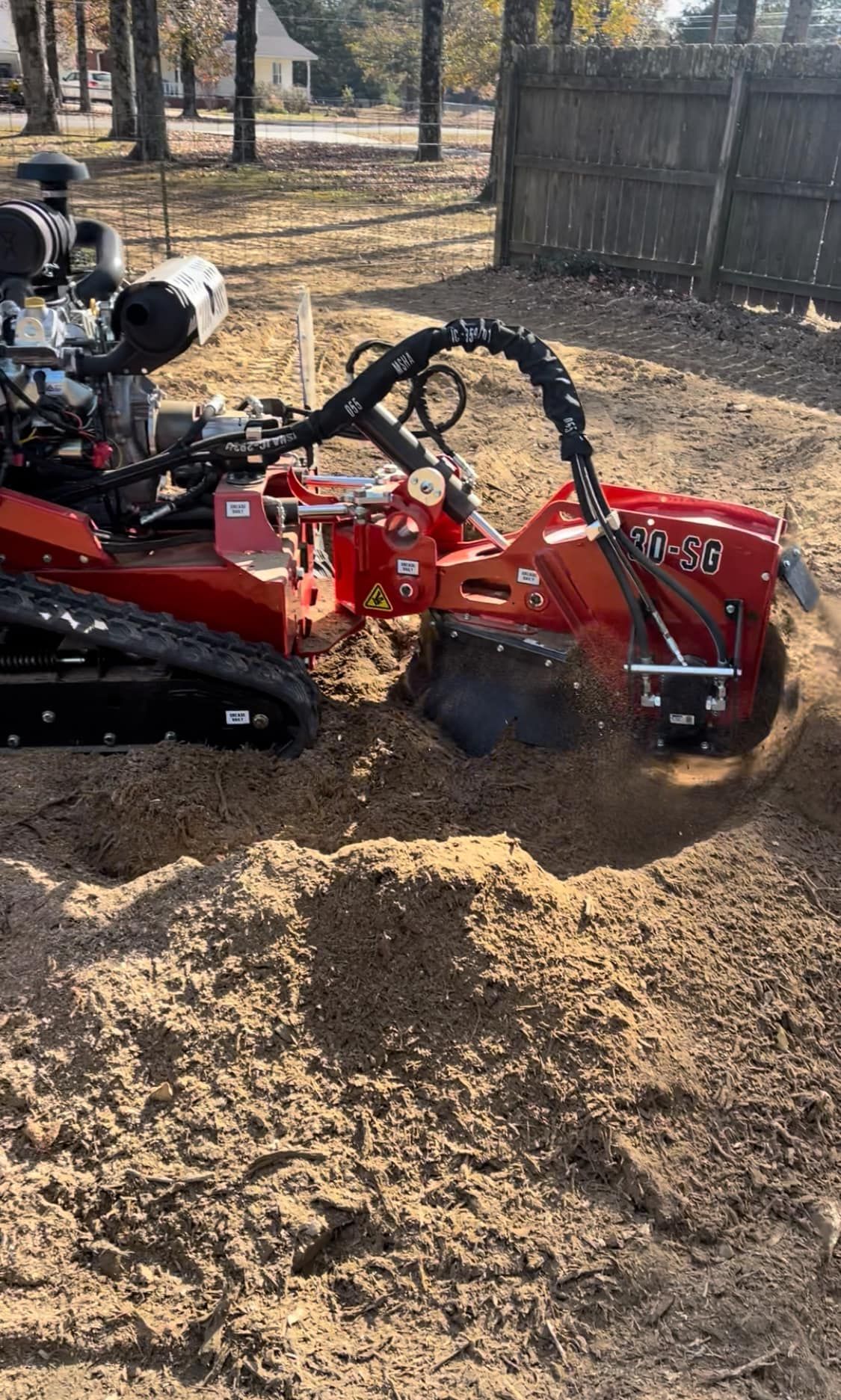 A red stump grinder with tracks is actively removing a tree stump in a yard, creating a pile of wood chips.