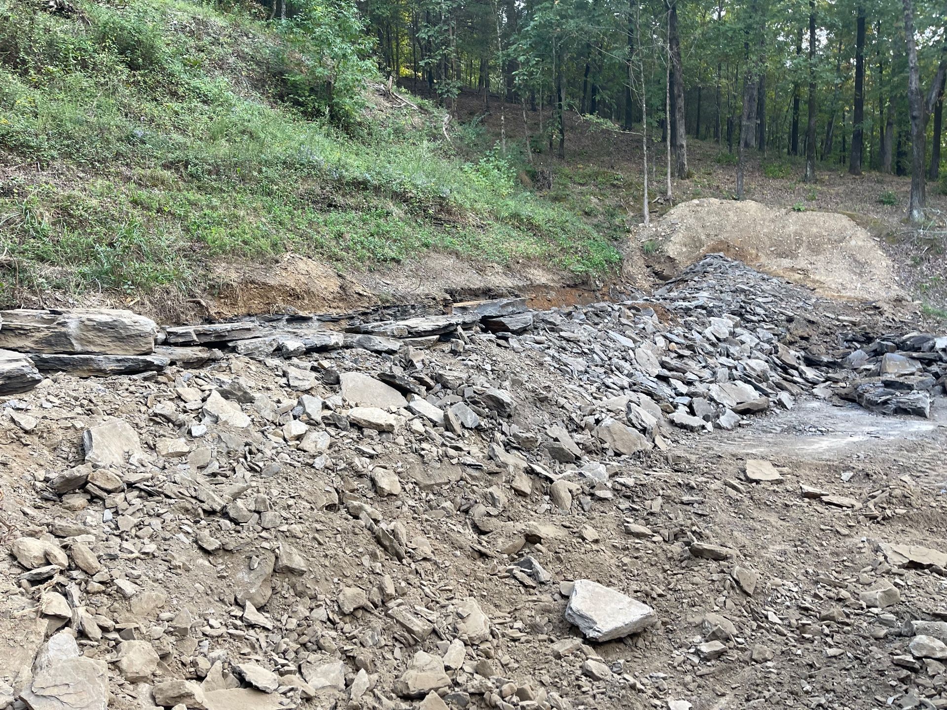 A rocky stream bed in a wooded area with exposed dirt banks and a small pile of excavated earth.
