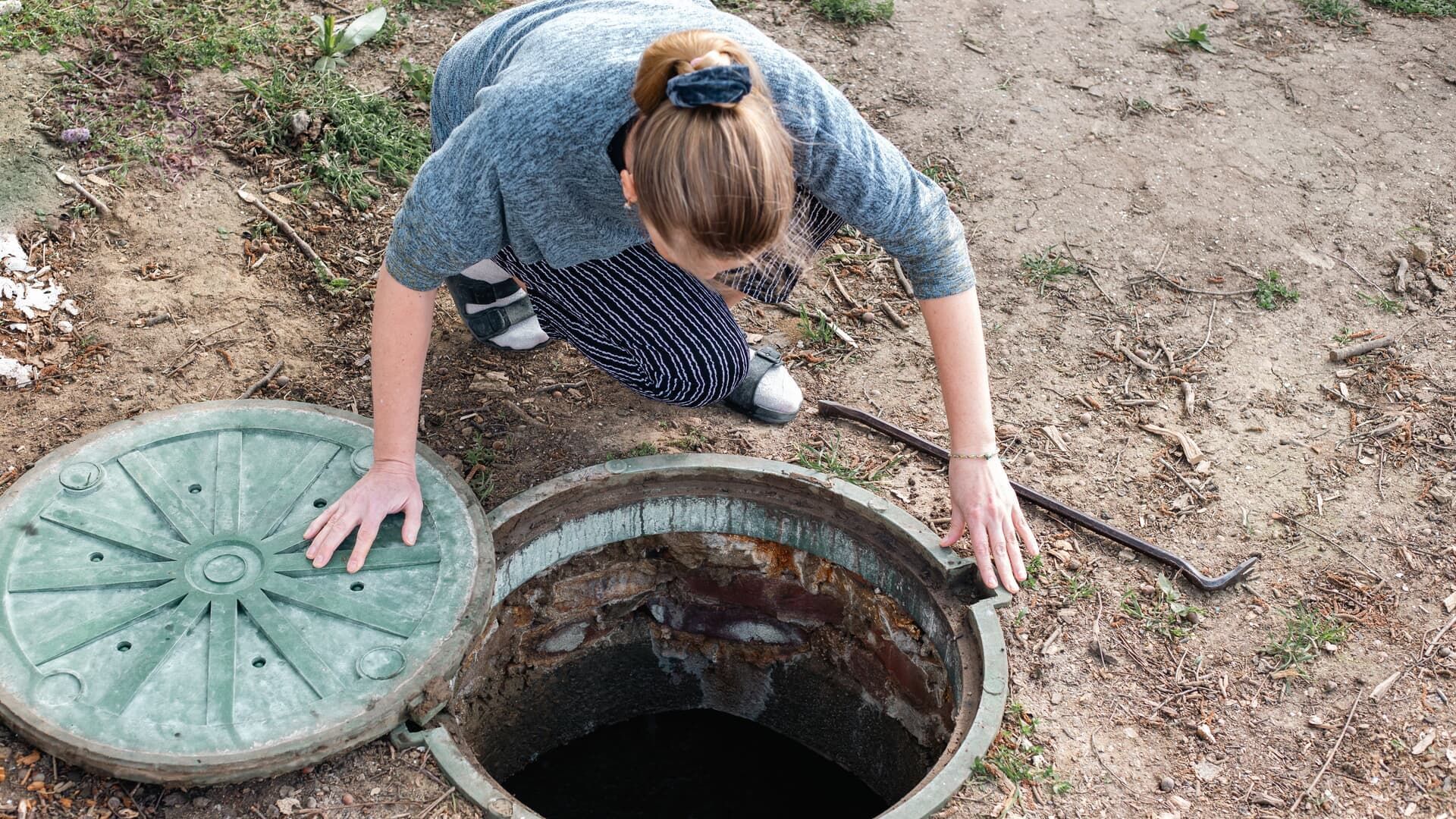 A person crouched over an open, circular septic tank cover in a grassy area, reaching toward the rim.