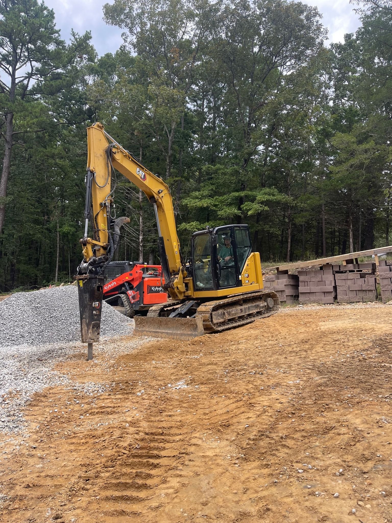 A yellow Caterpillar excavator with a hydraulic breaker attachment parked on a dirt lot near a retaining wall and trees.