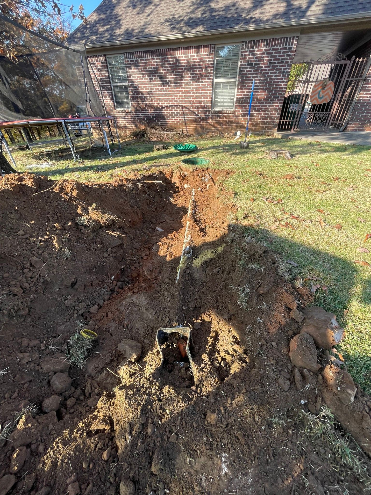 A deep, narrow trench dug into a residential lawn leads toward a brick house in the background.