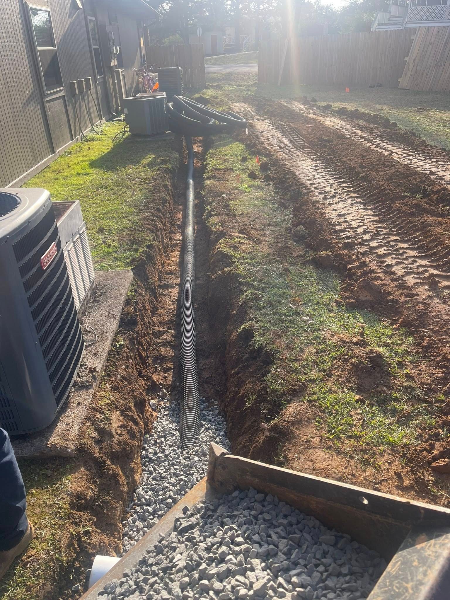 A trench with a drainage pipe and gravel base, located in a yard next to an air conditioning unit.