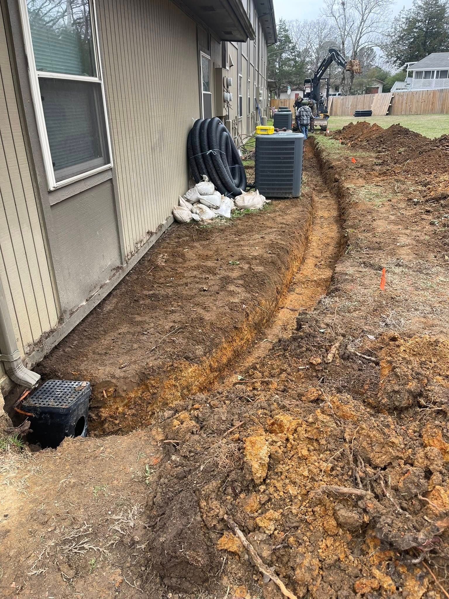 A trench dug along the side of a beige house with a drain basin, drainage pipes, and an AC unit nearby.