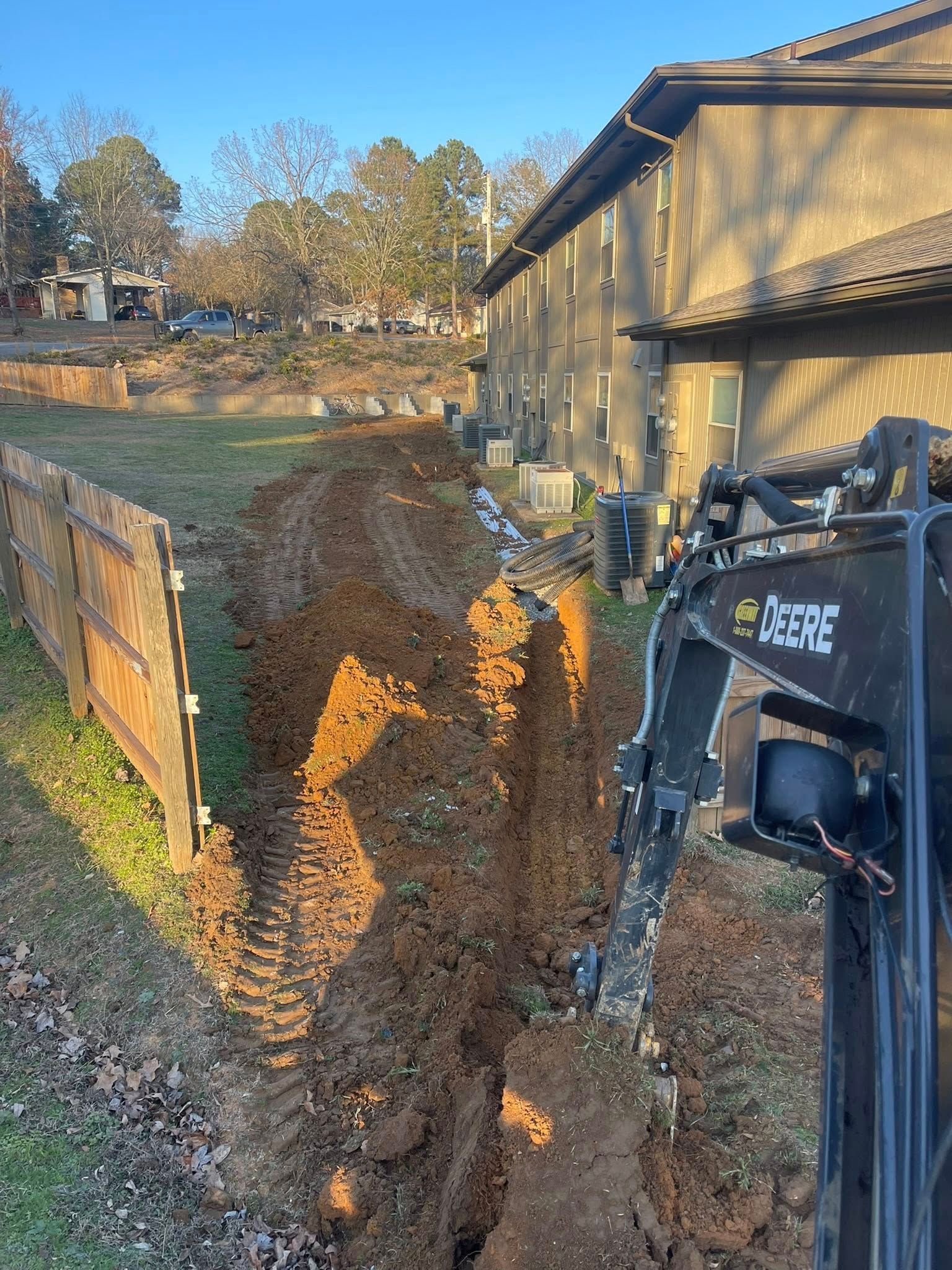 A John Deere excavator digs a trench in a dirt yard next to an apartment building and a wooden fence.