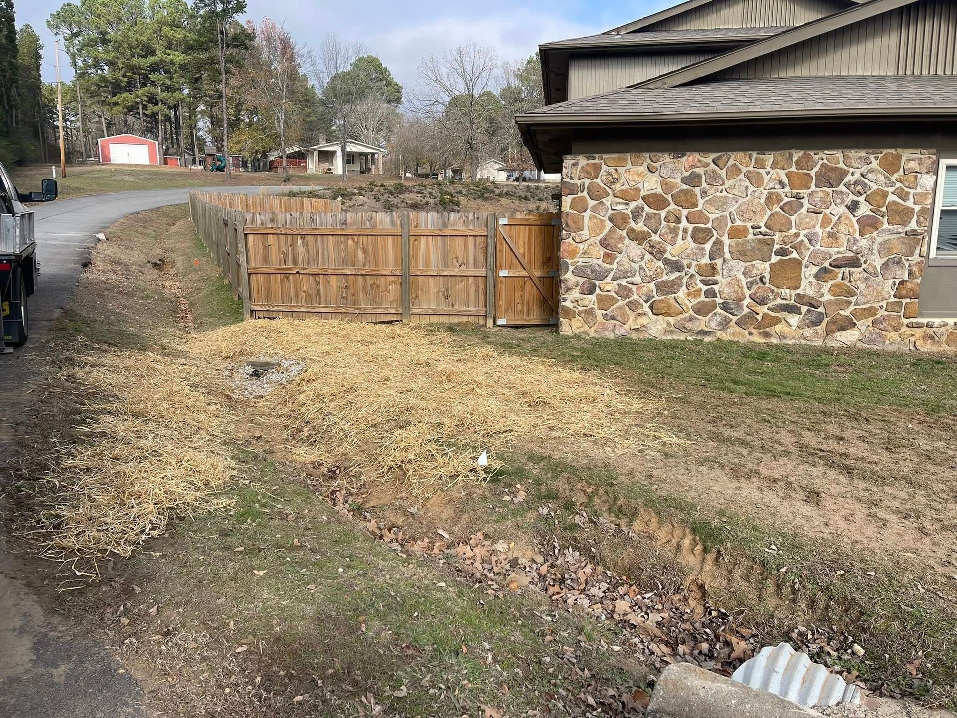 Wood chips cover the ground near a stone building and a wooden fence along a gravel road.