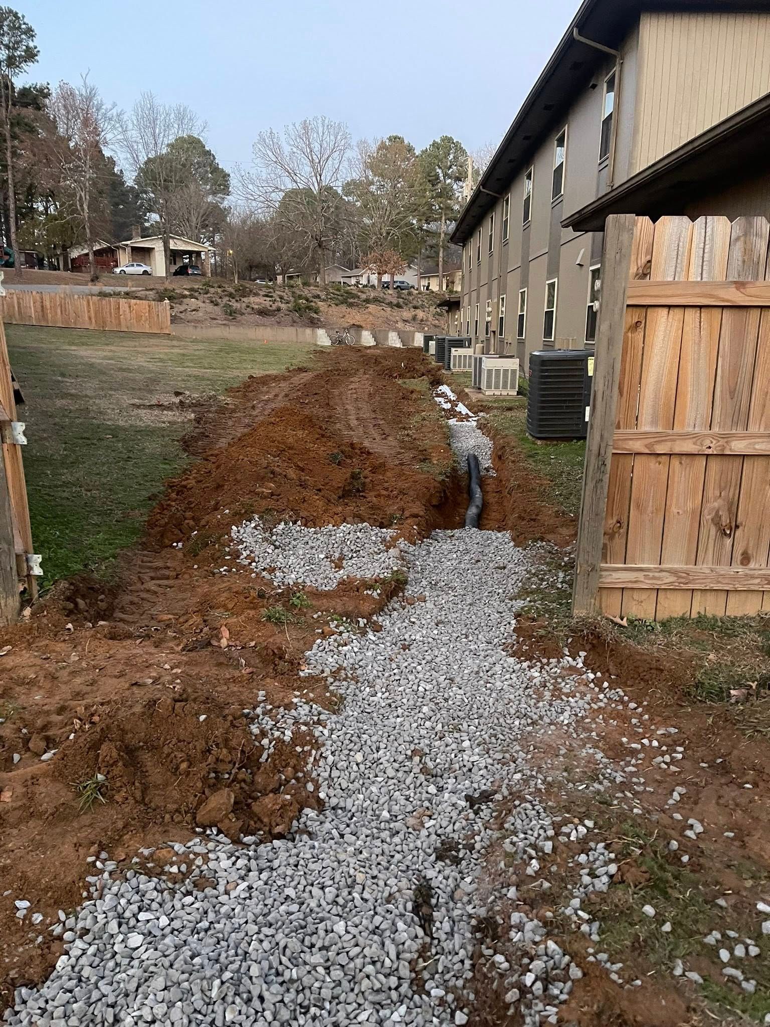 A gravel-filled drainage trench being installed in a yard next to an apartment building and a wooden fence.