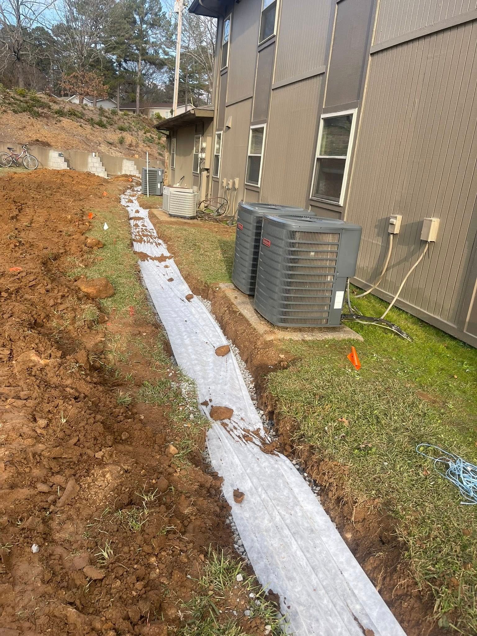 A trench filled with gravel runs along the side of a brown building next to two outdoor HVAC units and a dirt mound.