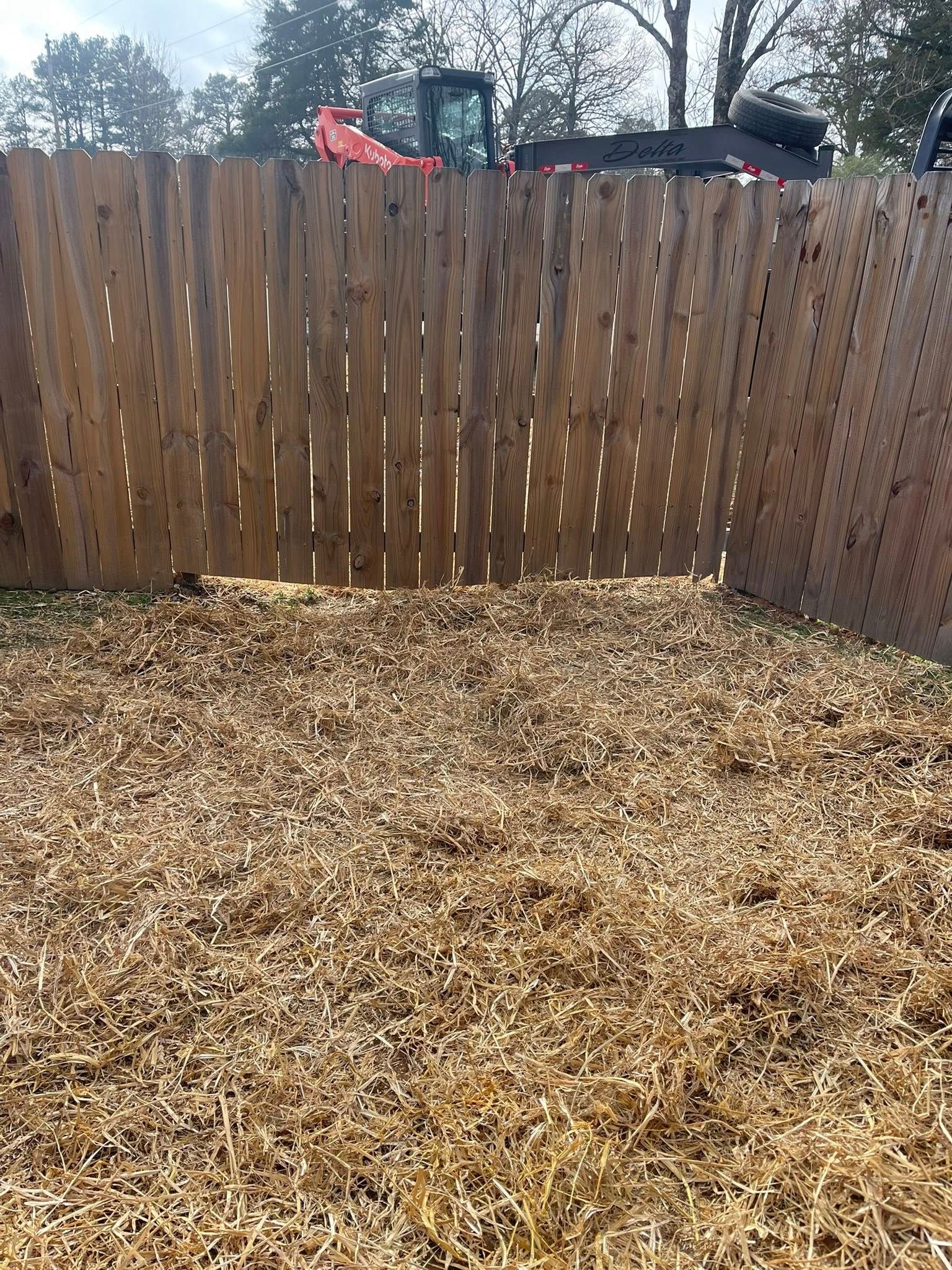 A wooden fence borders a backyard covered in fresh wood mulch, with construction equipment visible in the background.