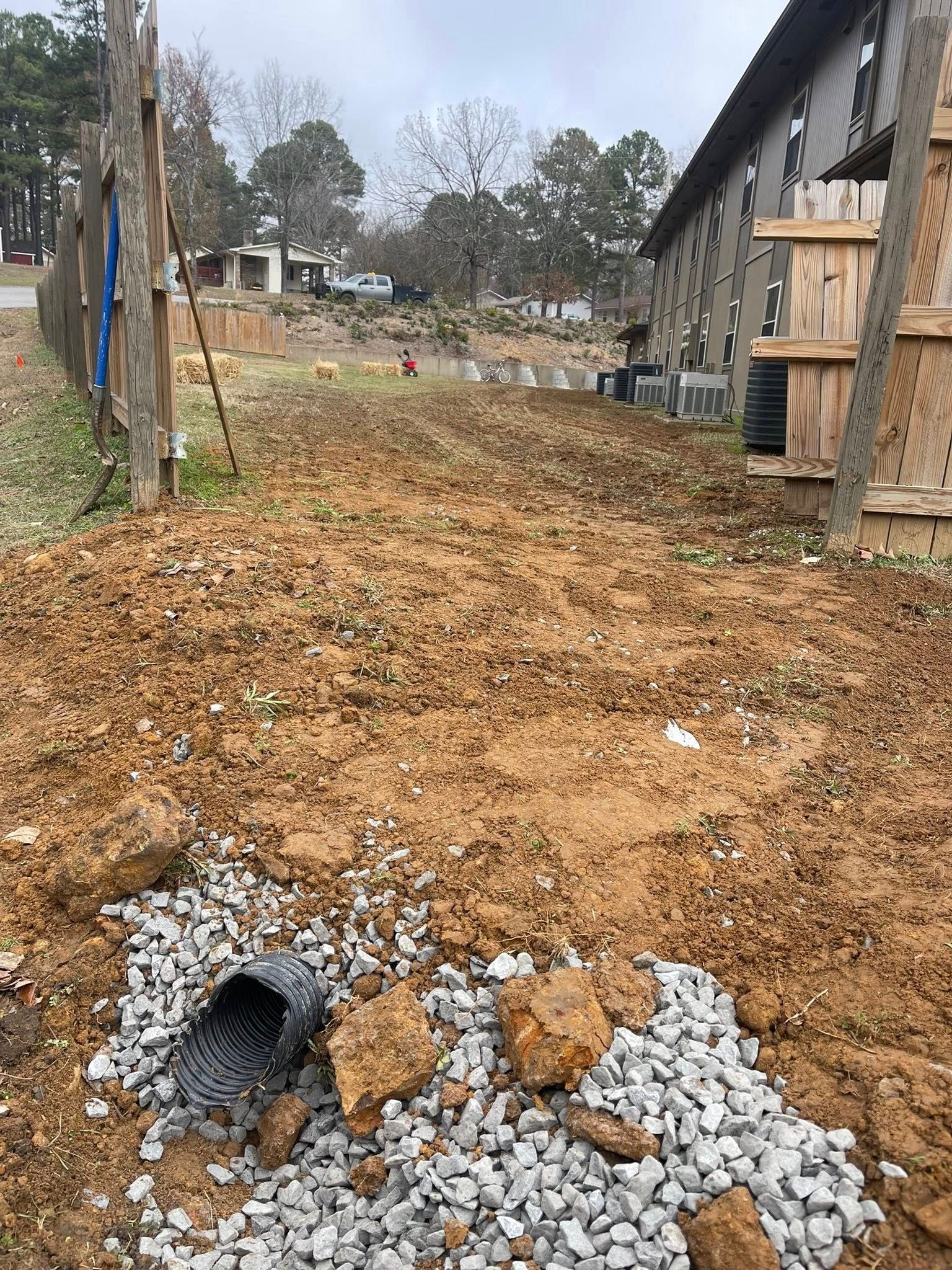 A drainage pipe end surrounded by loose gravel in a dirt lot beside a building under construction.