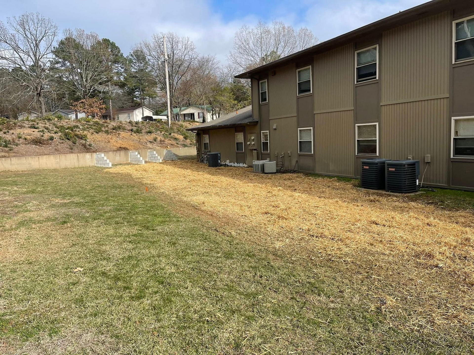 A brown, two-story apartment building with air conditioning units sits next to a lawn covered in wood chips.