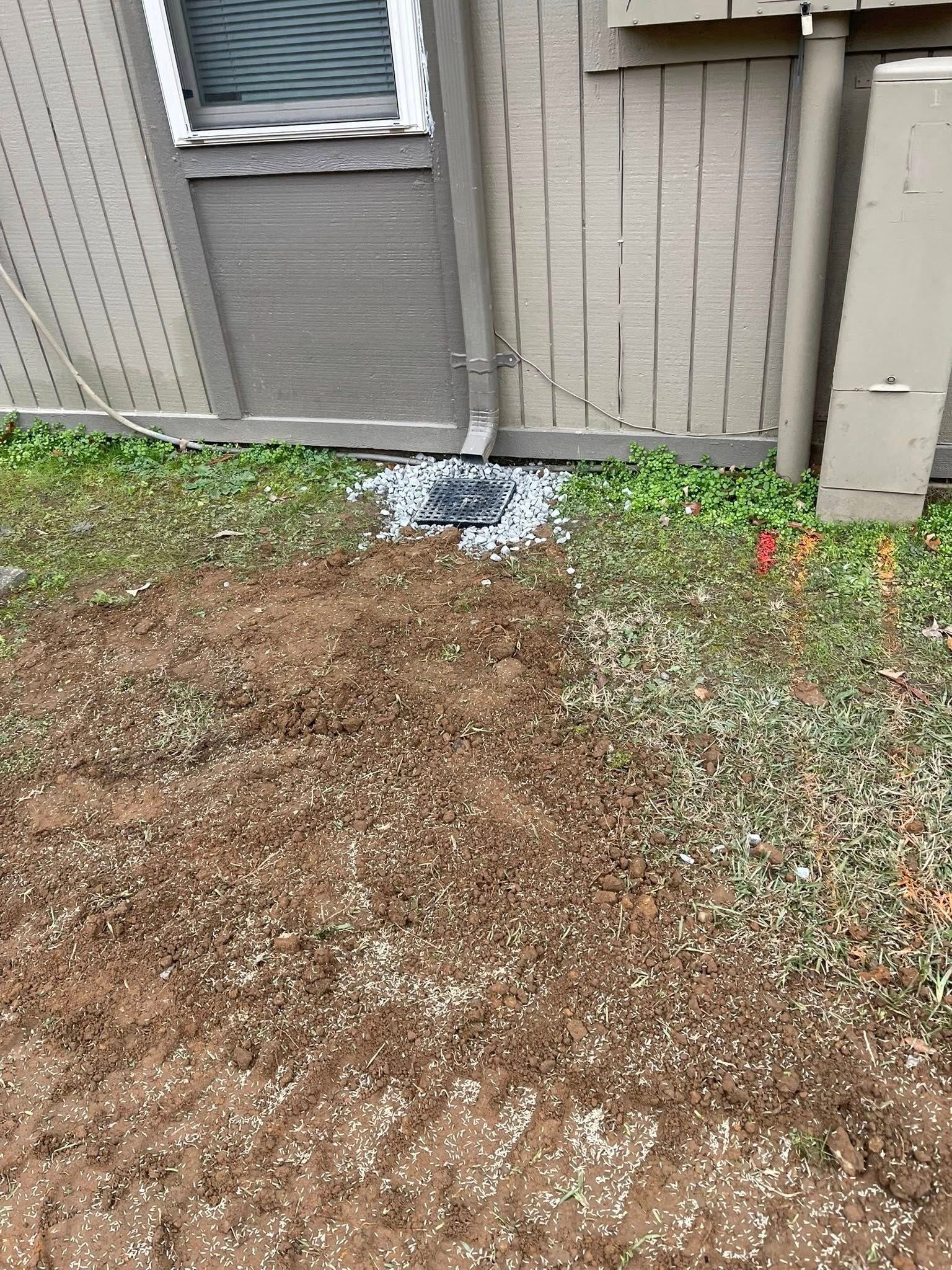 A downspout extends from the side of a tan house to a square drainage catch basin surrounded by white gravel.