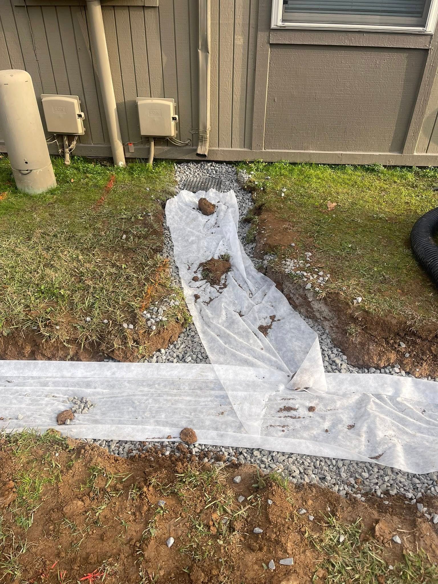 A gravel-filled trench lined with white landscape fabric sits in a grassy yard next to a building foundation.