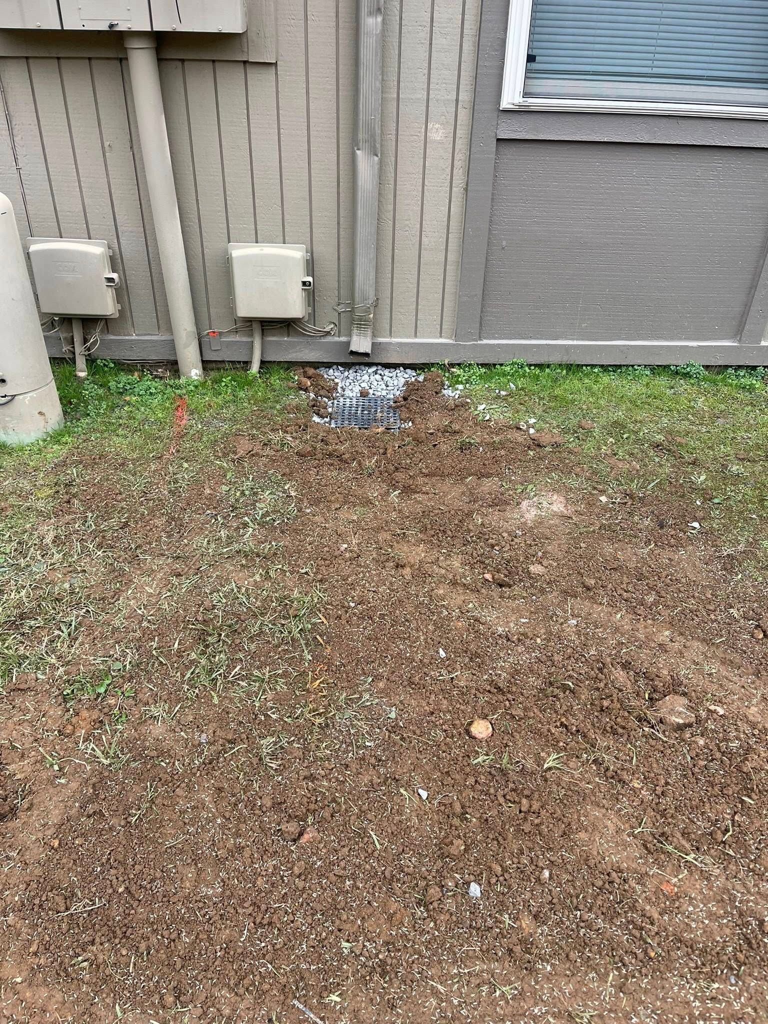 A downspout leads into a square drainage grate located in a patch of dirt and sparse grass against a tan exterior wall.