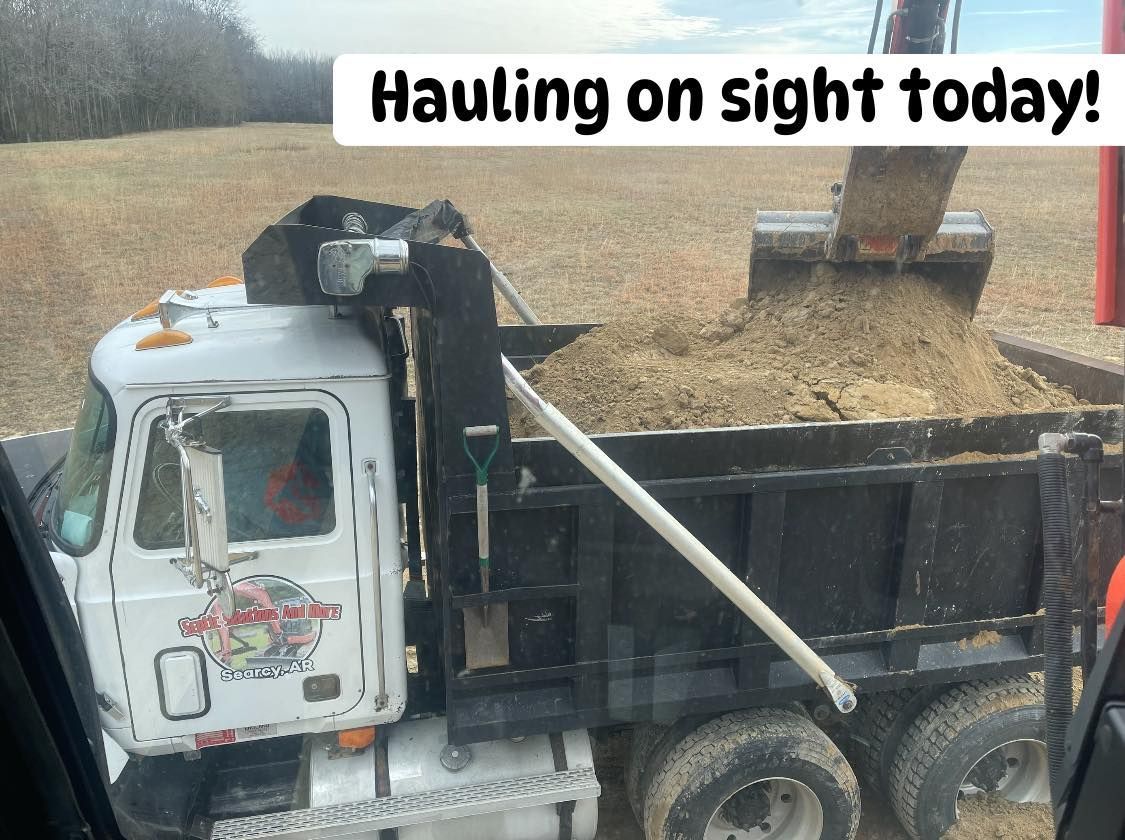 An excavator loads dirt into the back of a white dump truck parked in an open field, with the text 
