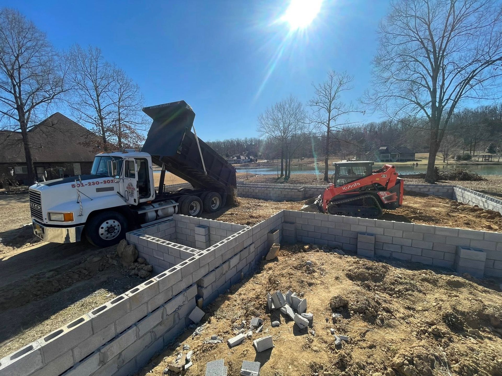 A white dump truck unloads dirt into a cinder block building foundation while a small orange skid-steer works nearby.