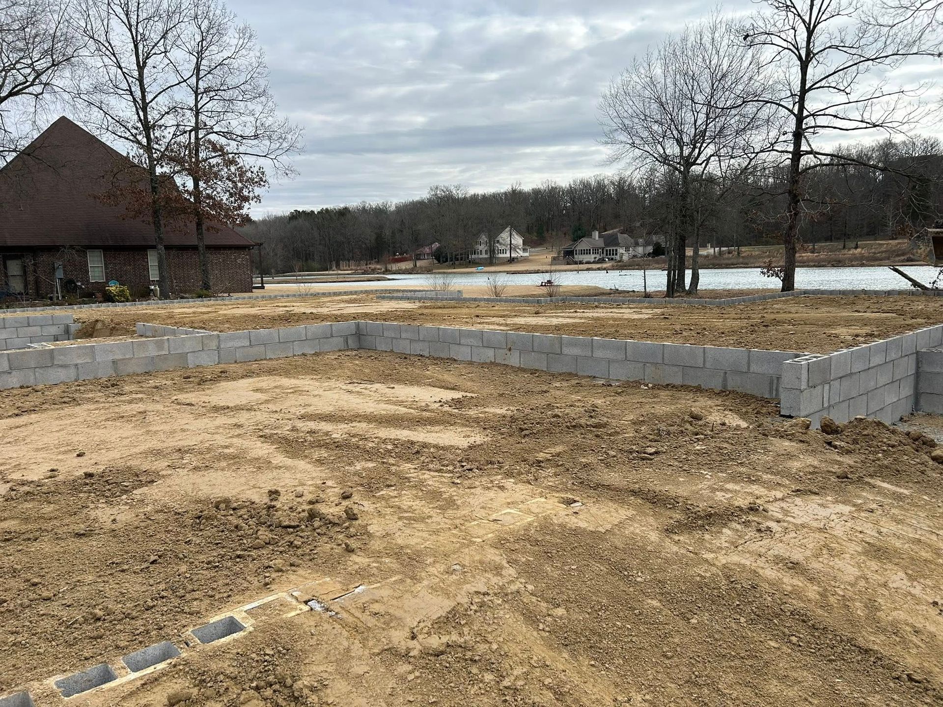 A residential construction site with a concrete block foundation set in a dirt yard, near a house and trees under a cloudy sky.