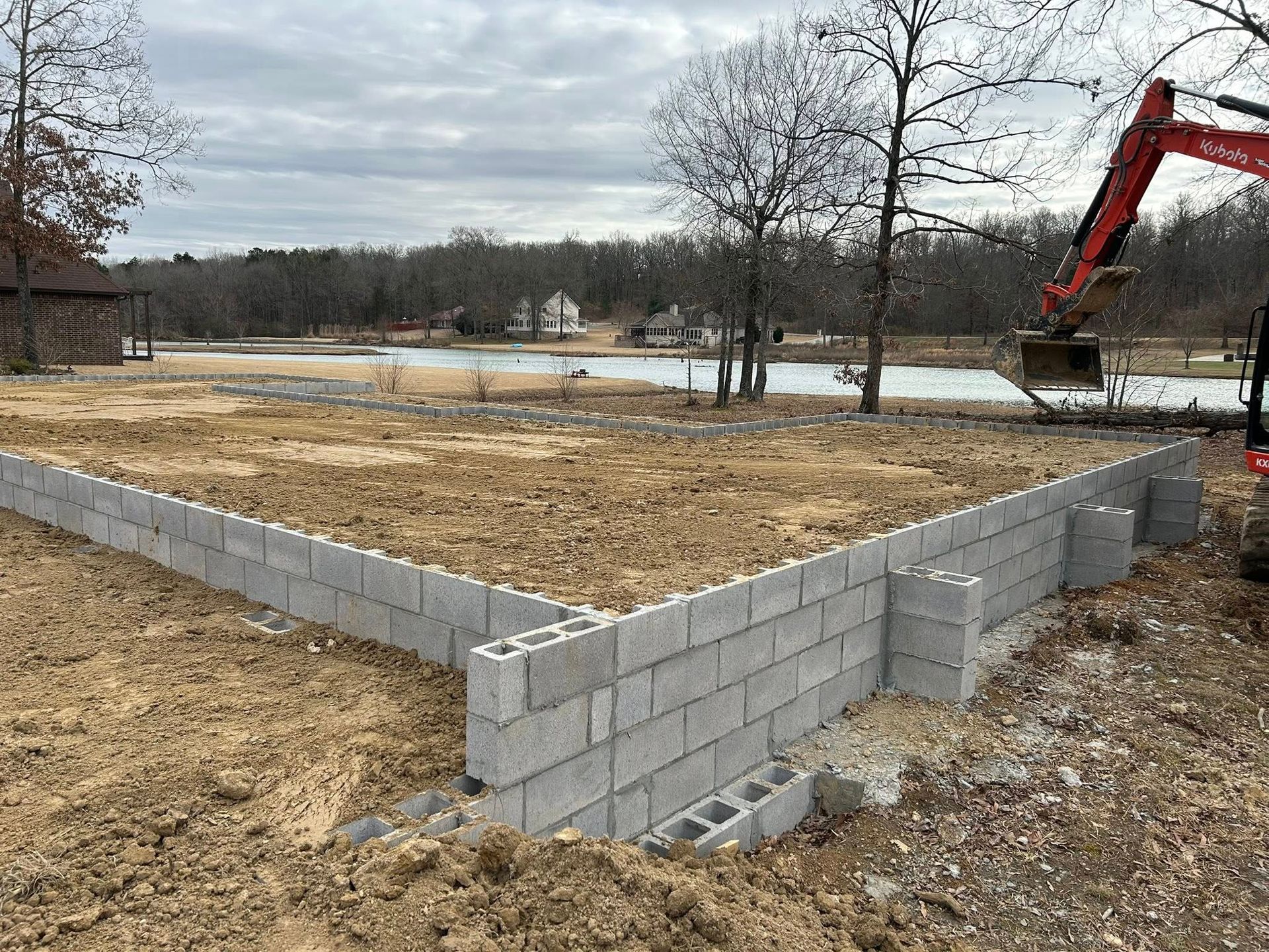 A concrete block foundation under construction at a rural site near a lake, with an excavator visible in the background.
