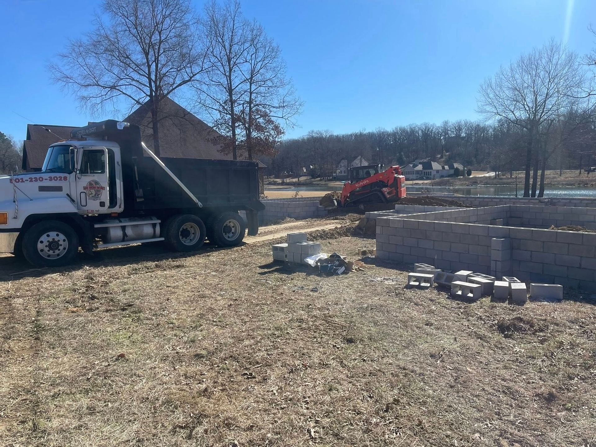 A white dump truck parked next to a stone retaining wall under construction, with a small orange skid steer in the distance.