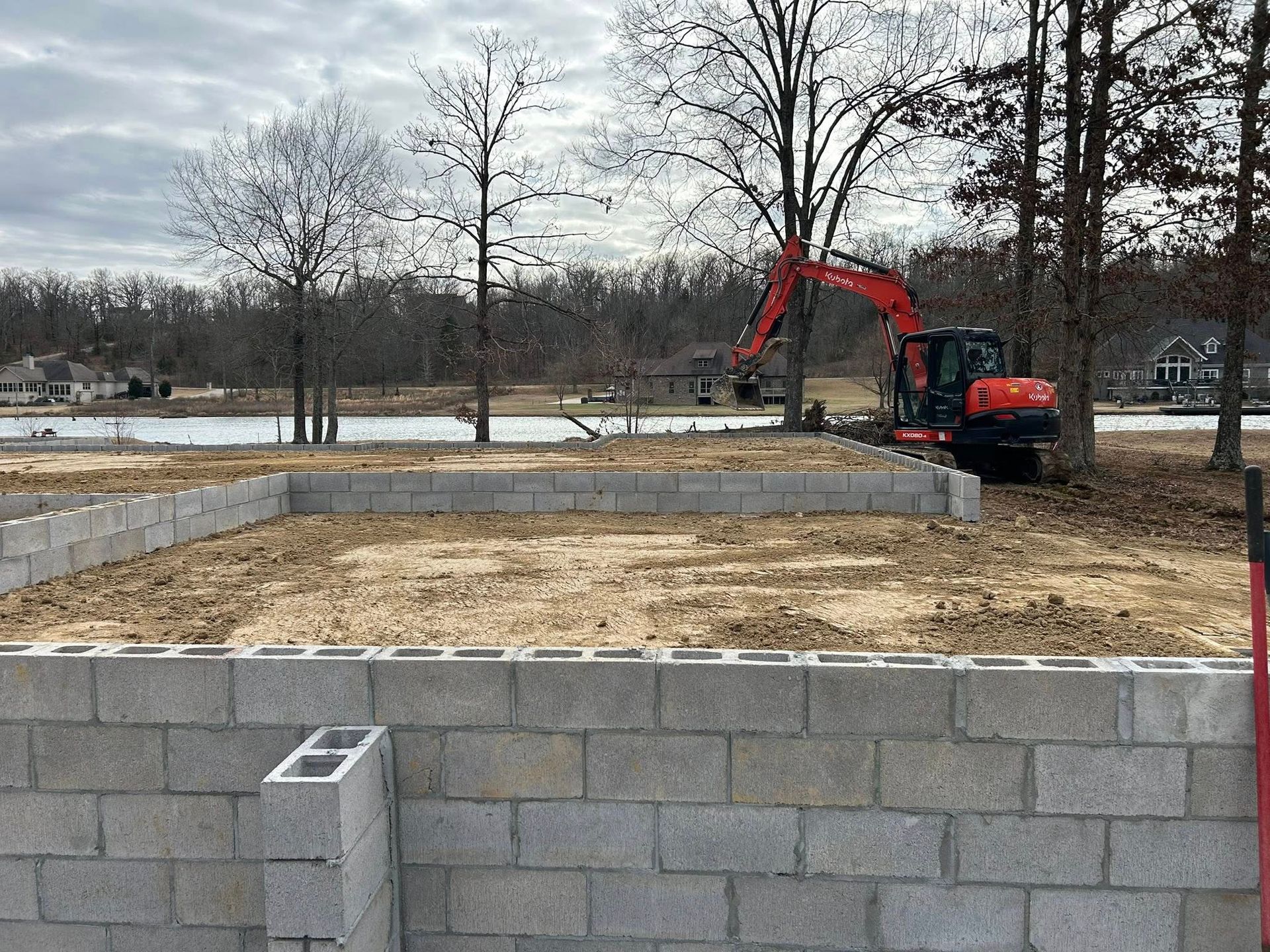 An orange excavator on a construction site near a pond, behind a partially built cinder block wall.
